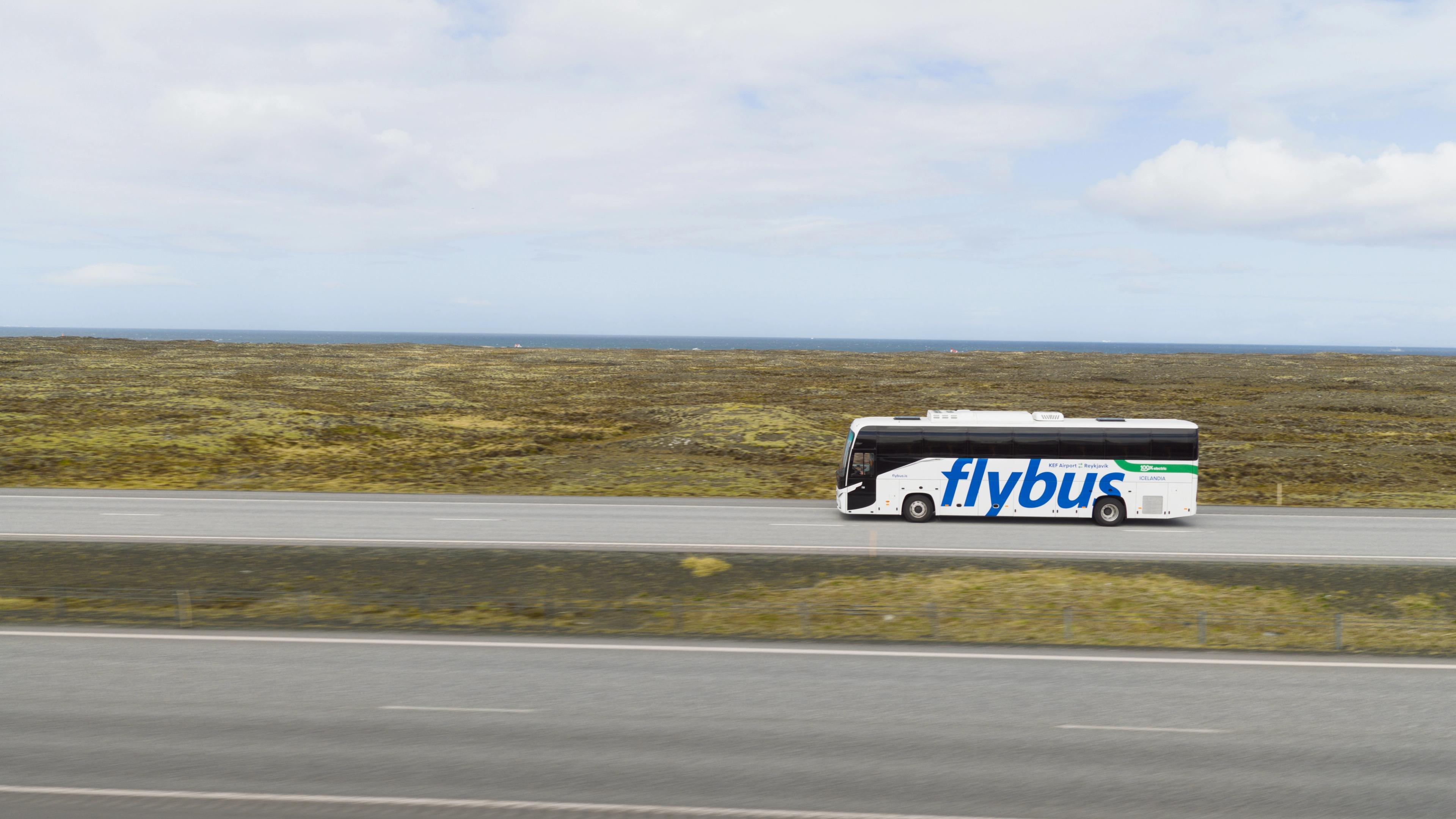 A white Flybus drives on a highway through a mossy landscape with the sea and cloudy sky in the background.