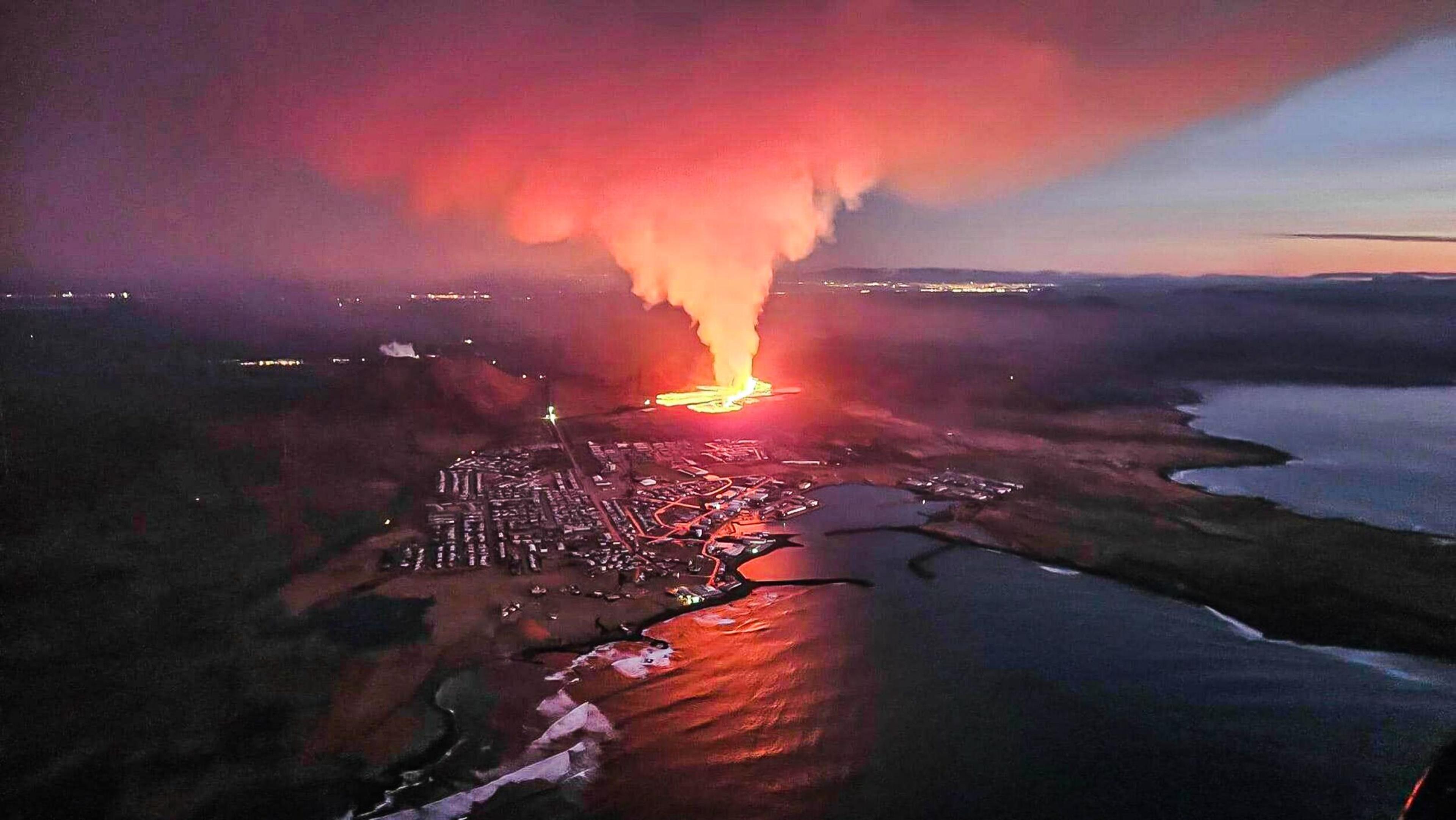Aerial view of Grindavík's volcanic eruption at dusk, with red-orange lava and smoke against twilight sky.