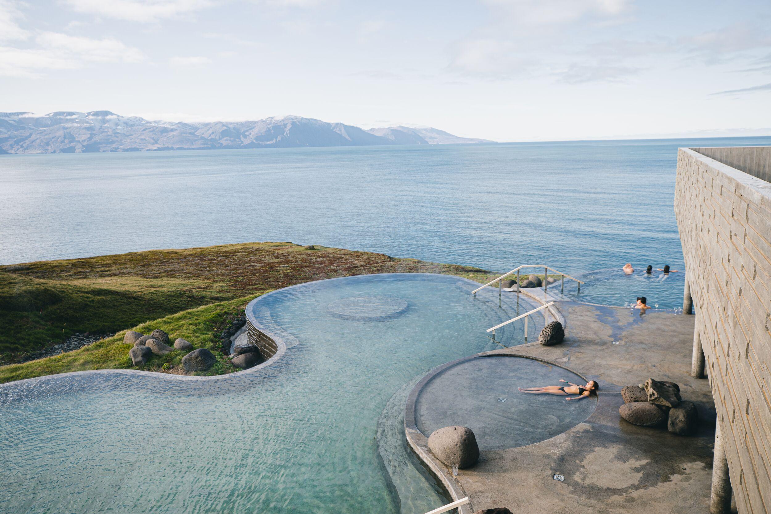 Outdoor geothermal pools on a grassy cliff overlooking a vast ocean and distant mountains, with people relaxing in the water.