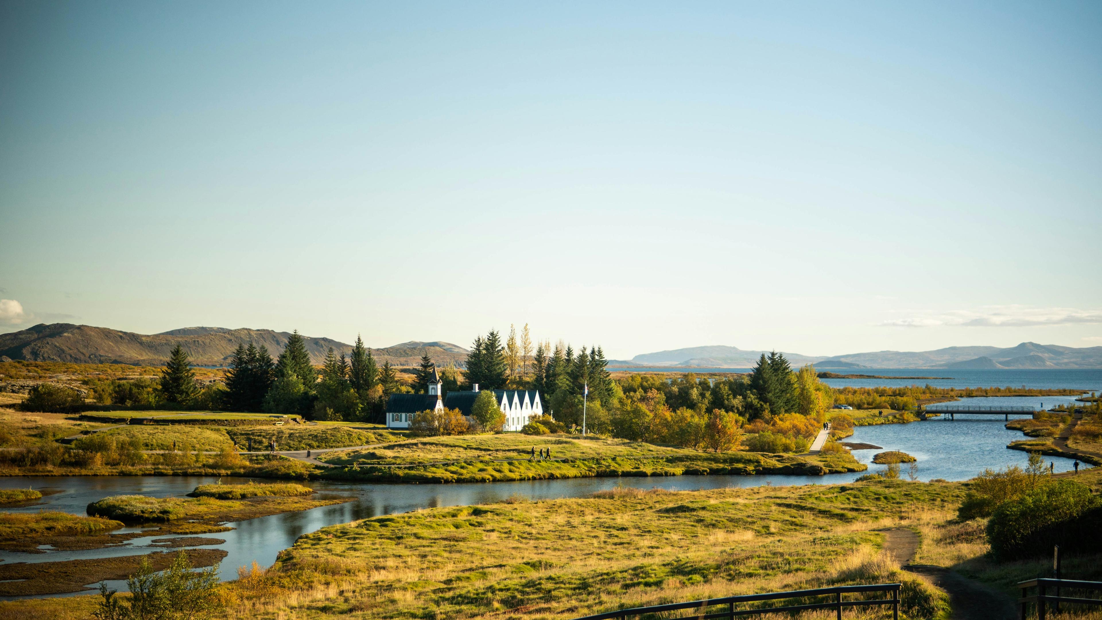 A serene landscape bathed in warm sunlight, featuring a small settlement with traditional buildings near a river, nestled within the vast, open plains and gentle hills, reminiscent of Þingvellir (Thingvellir) National Park in Iceland.