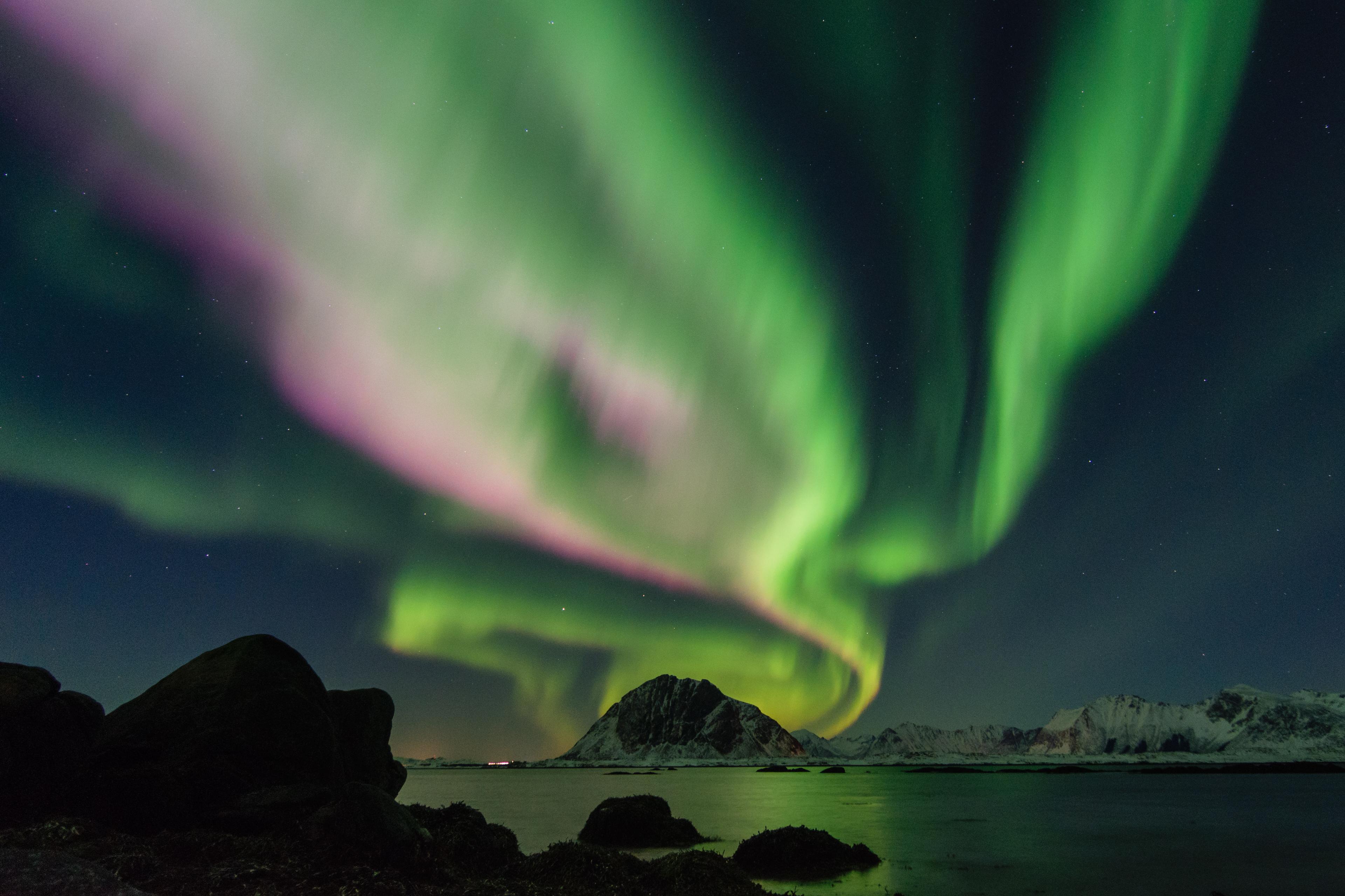 A stunning aurora borealis swirls in the sky, with shades of green and pink, over a serene coastal landscape with mountains in the background.