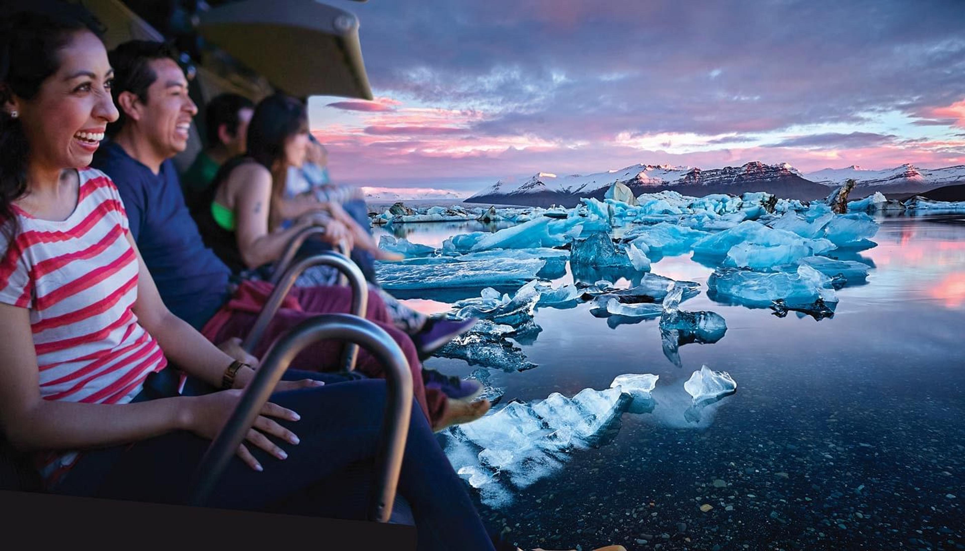 Smiling people in an immersive simulator view a glacial lagoon with icebergs and mountains under a colorful sunset sky.