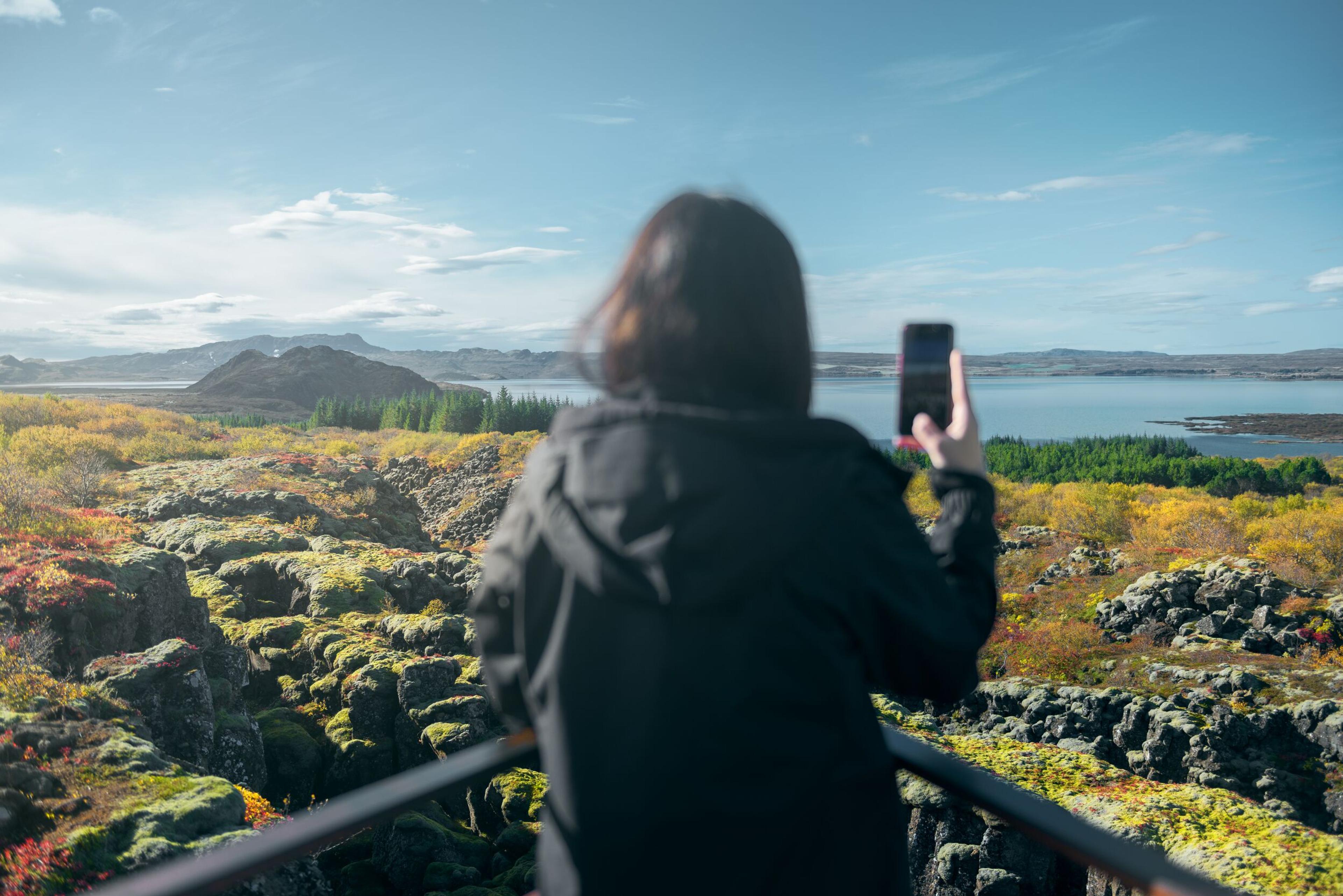 A person with their back to the camera takes a photo with a smartphone, capturing a landscape featuring autumn-hued vegetation and a lake, with mountains in the distance under a partly cloudy sky.