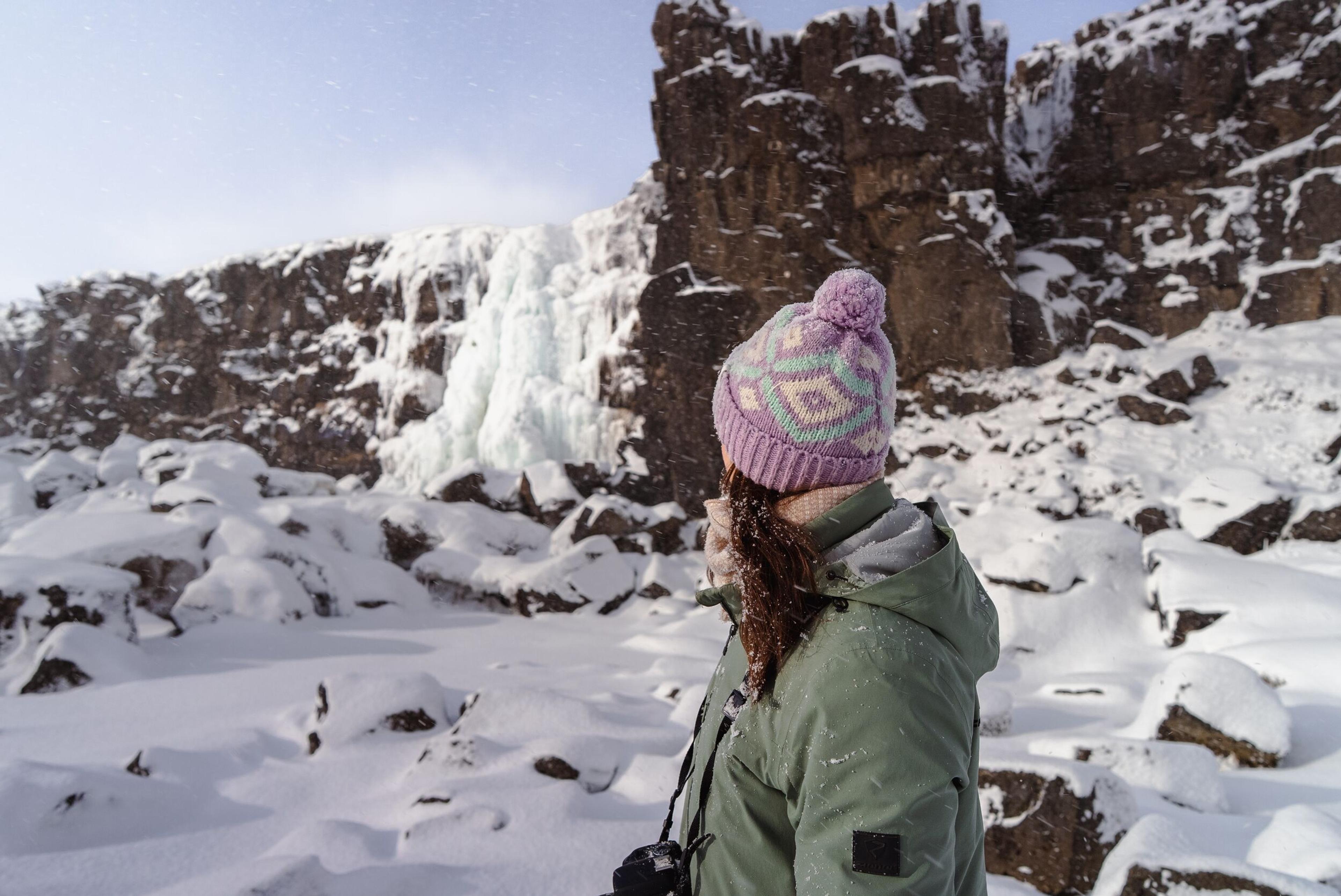 A person in a purple hat and green jacket looks at a large frozen waterfall against a snowy, rocky landscape.