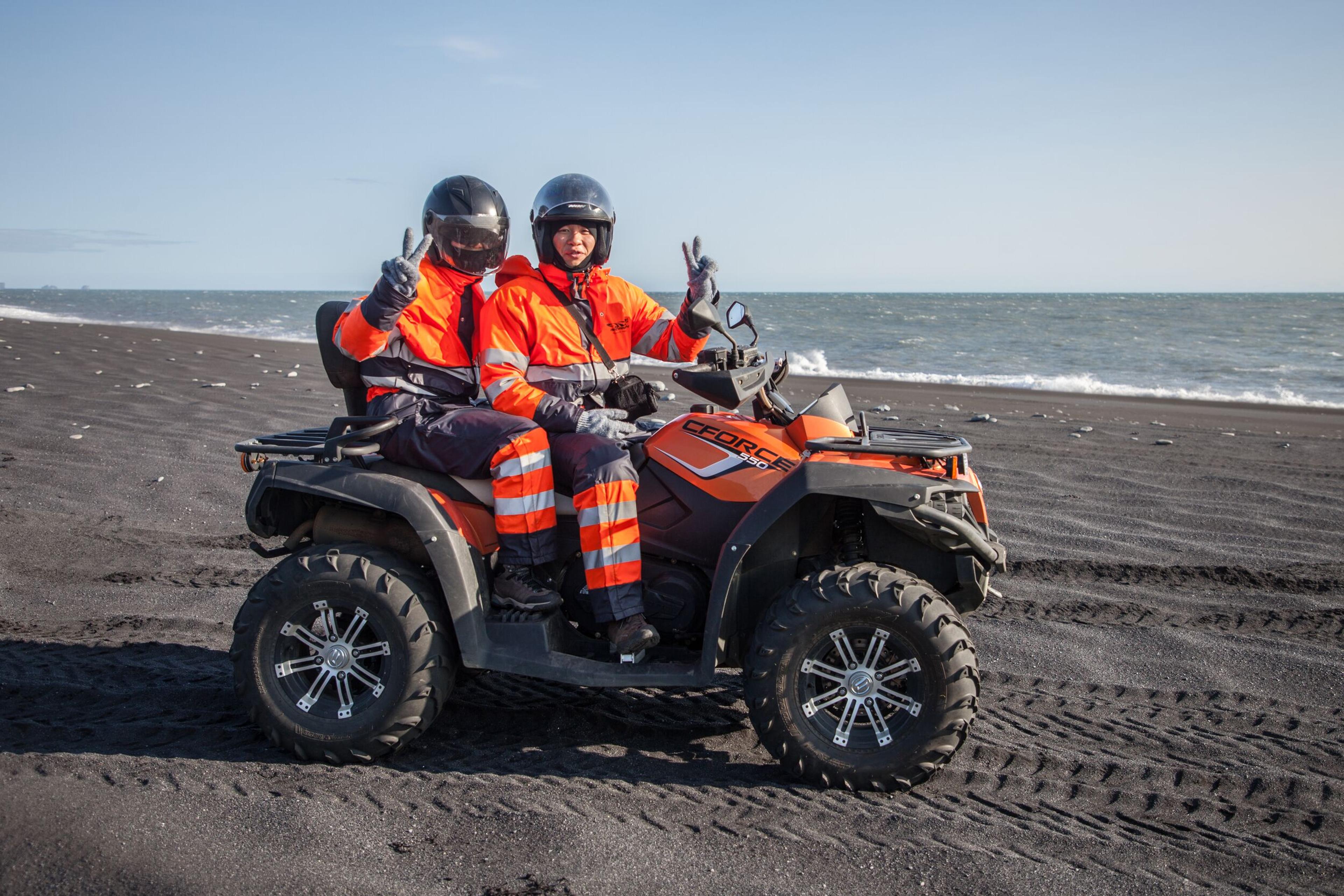 Two people in helmets and orange suits on an ATV give peace signs on a black sand beach with the ocean in the background.