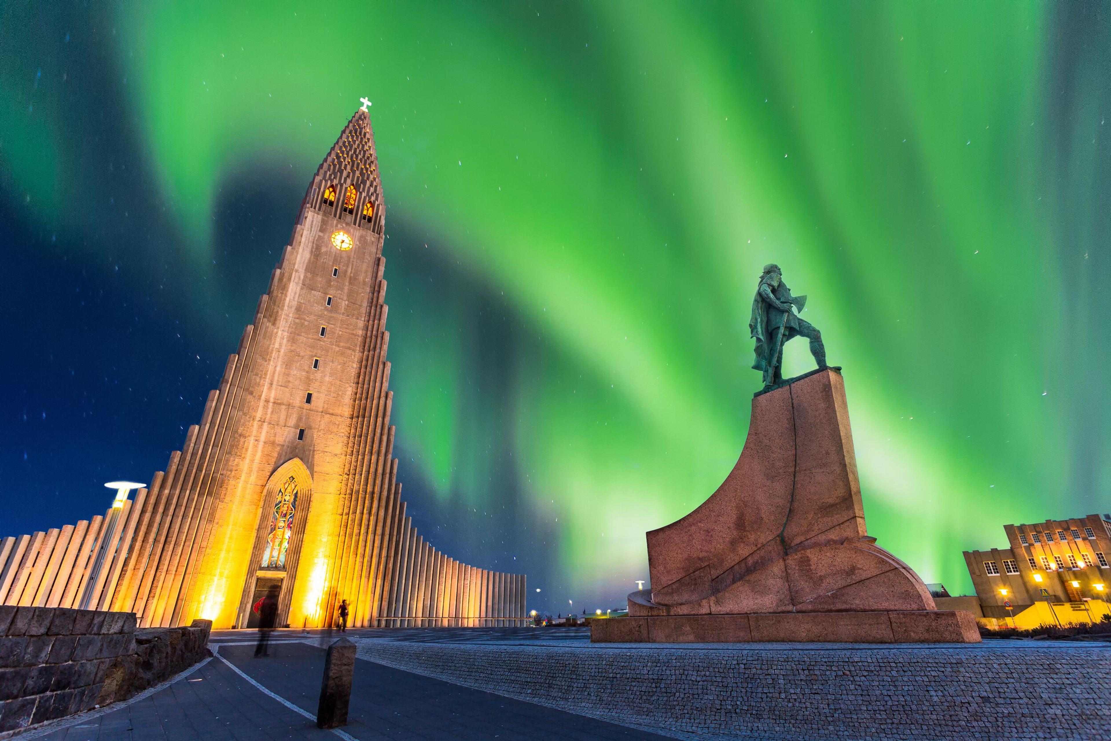 The Hallgrímskirkja church in Reykjavik, lit up at night, with the Northern Lights dancing in the sky above. A statue of Leif Erikson stands in the foreground