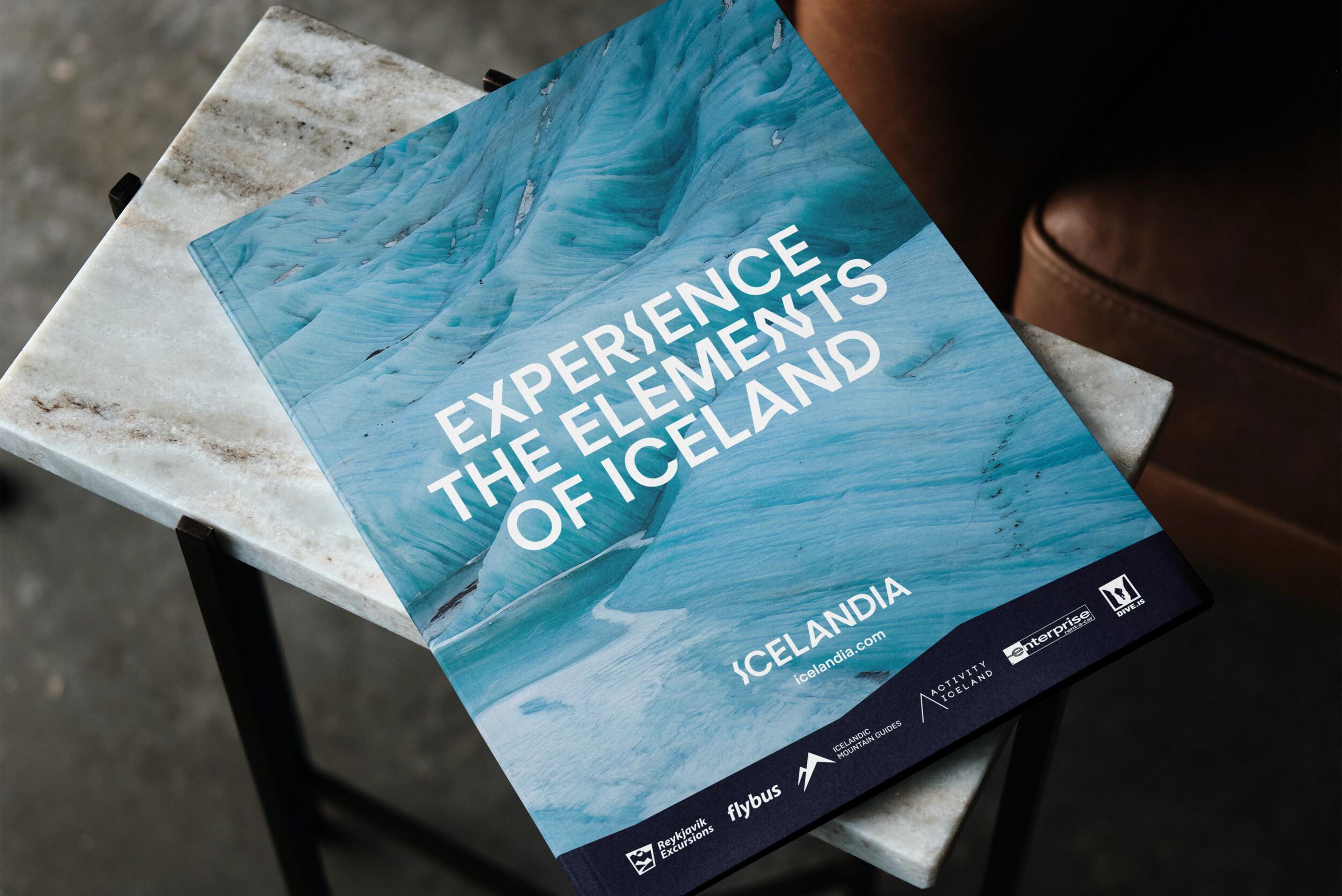 Cover of Icelandia brochure titled 'Experience the Elements of Iceland' with a glacier background, placed on a marble table."