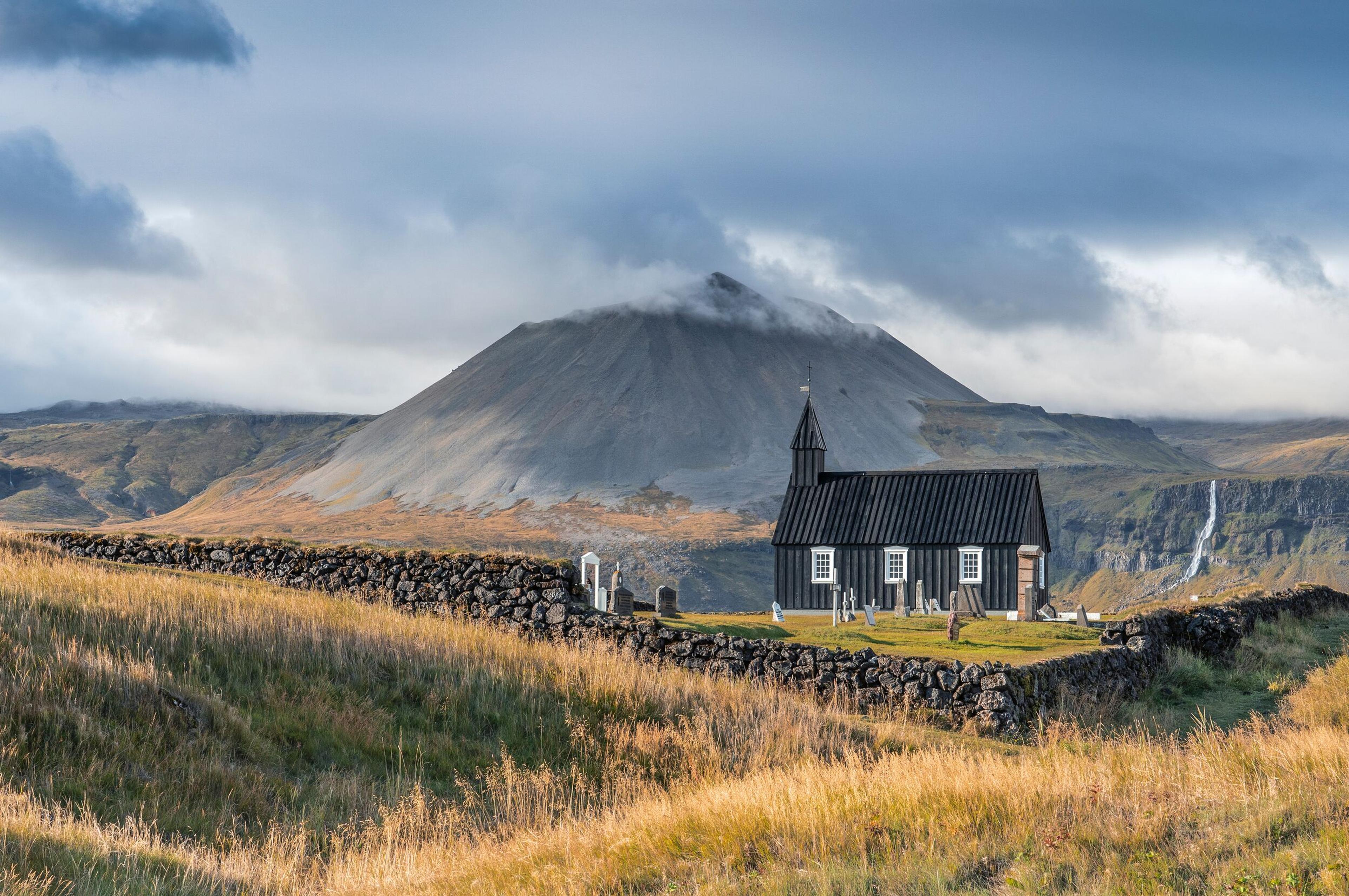 A quaint black church with a pointed roof, surrounded by a stone wall, stands in a grassy field with a majestic, mist-covered mountain in the background.