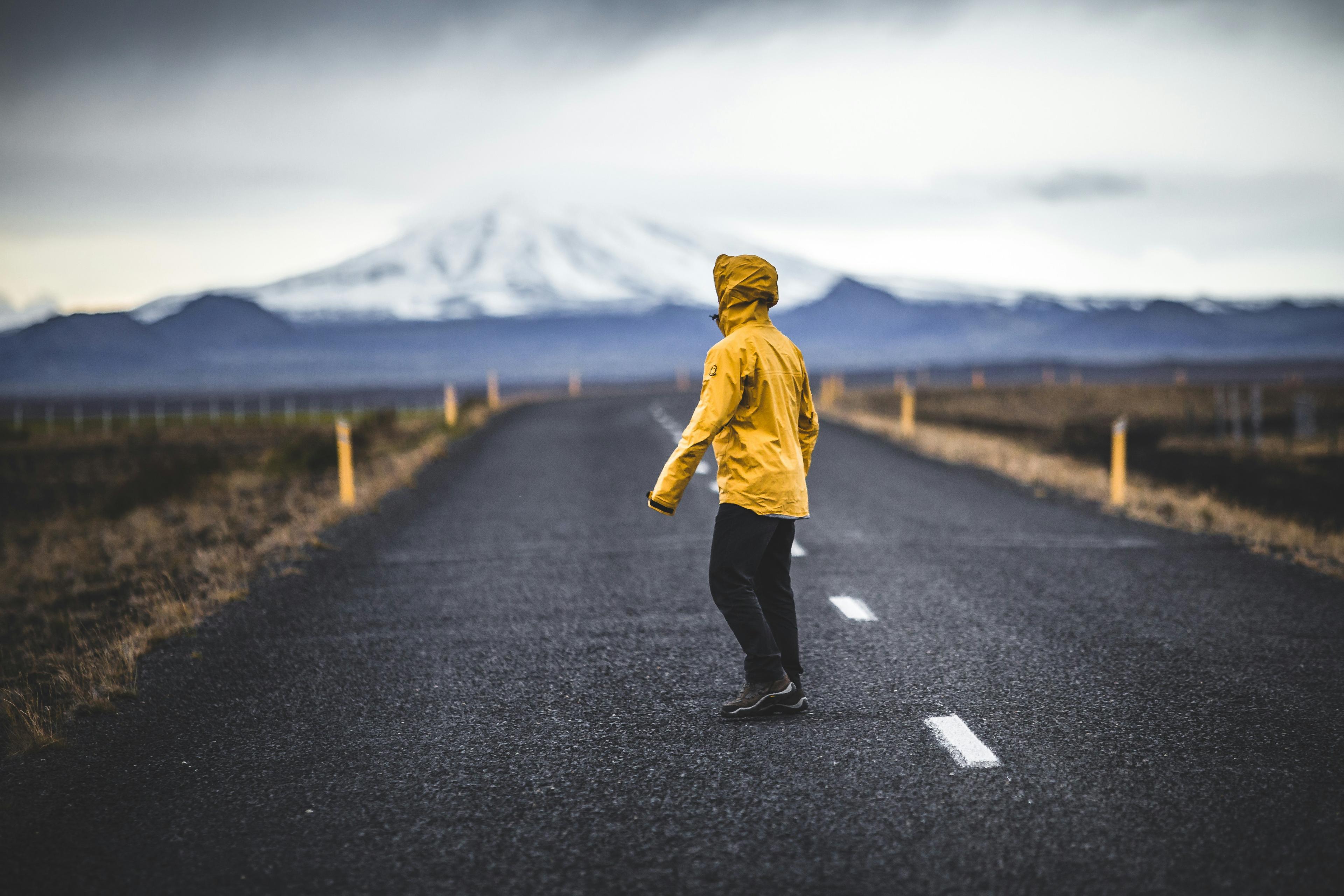 A person in a yellow rain jacket walking down an empty road towards a snow-capped mountain in Iceland.