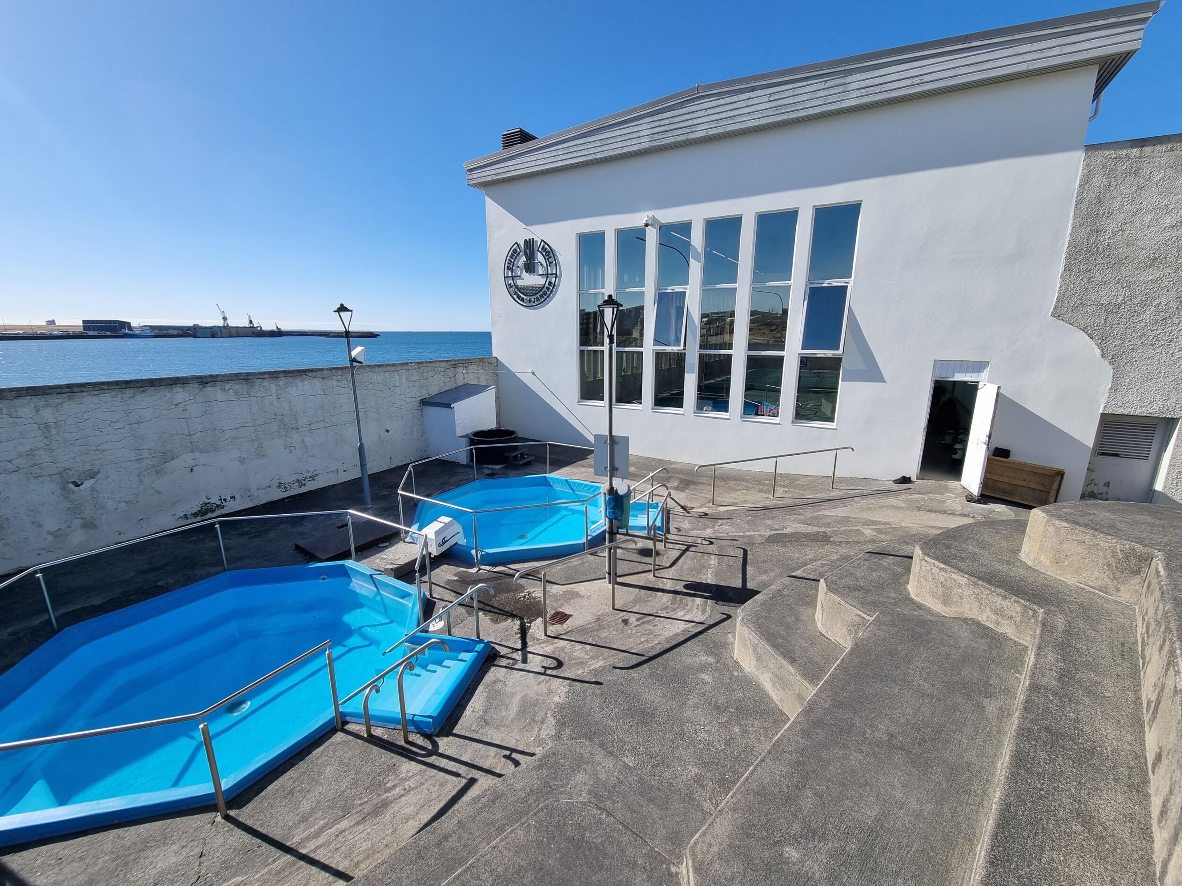 Two empty blue pools on a concrete deck next to a modern white building with large windows, overlooking a harbor under a clear blue sky.