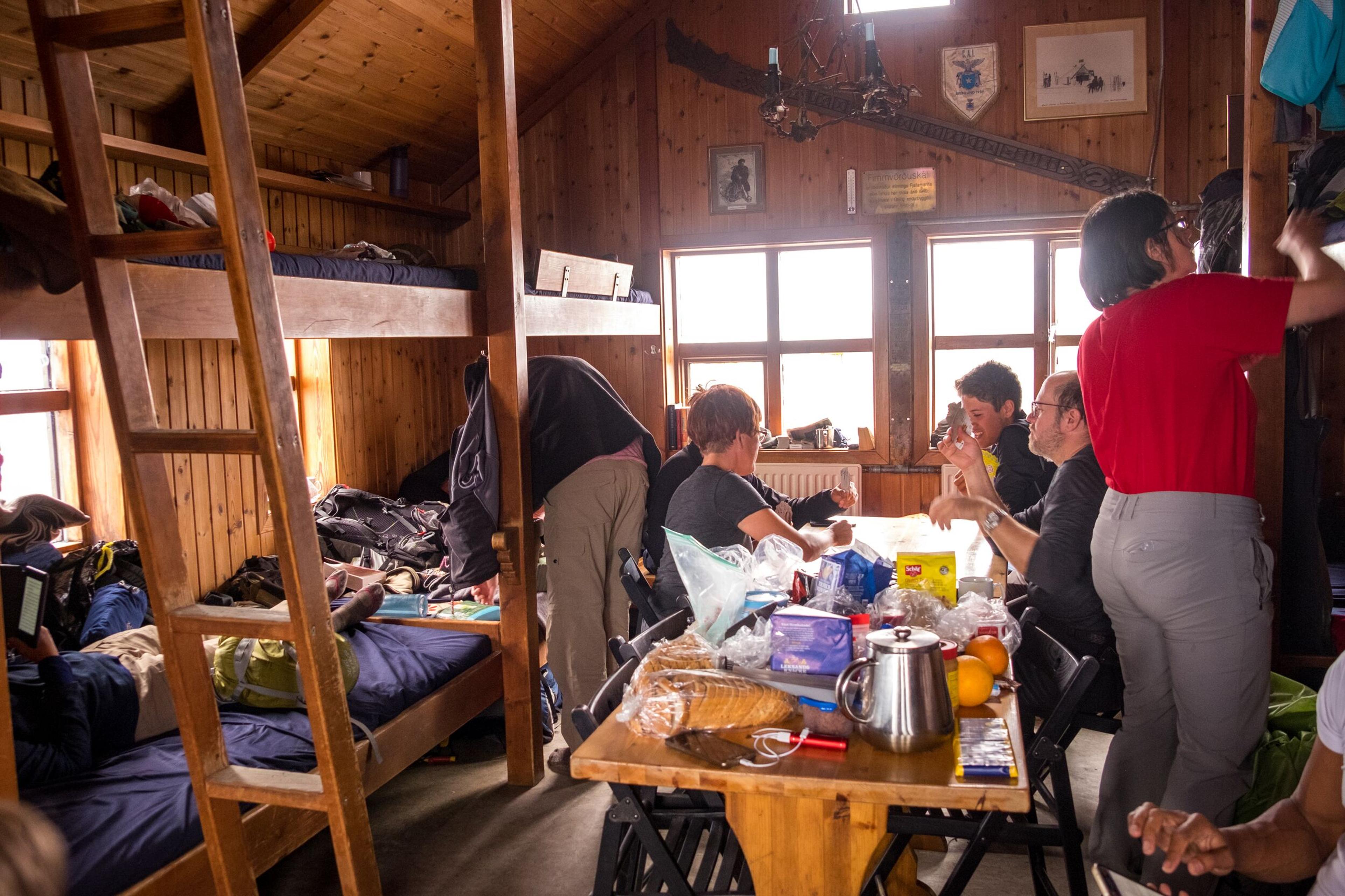 Hikers relaxing and eating inside a cozy mountain hut along the Laugavegur trail, with wooden bunk beds and a shared table.