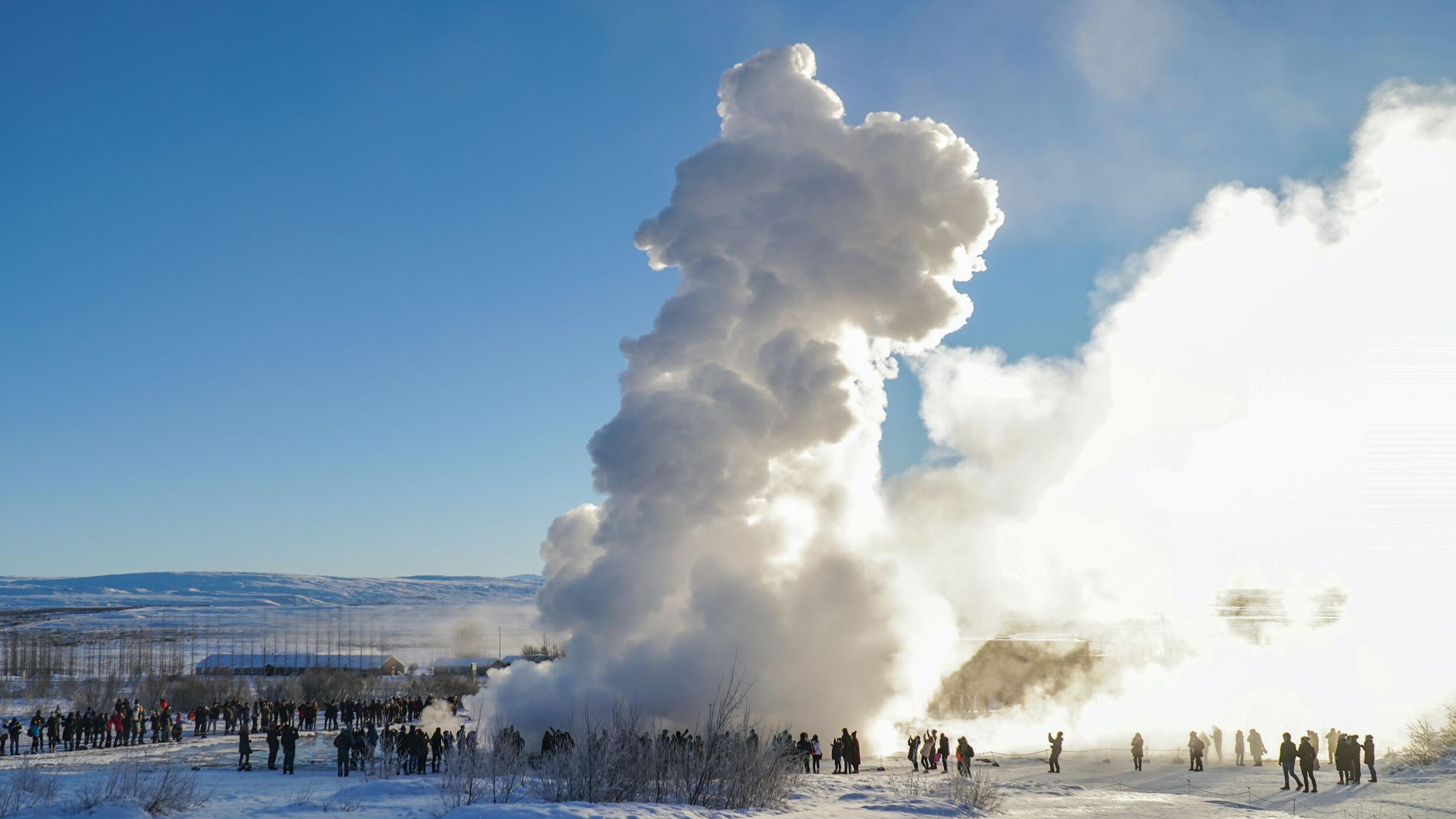 A geyser erupts with a large plume of steam, watched by a crowd in a snowy landscape under a blue sky.