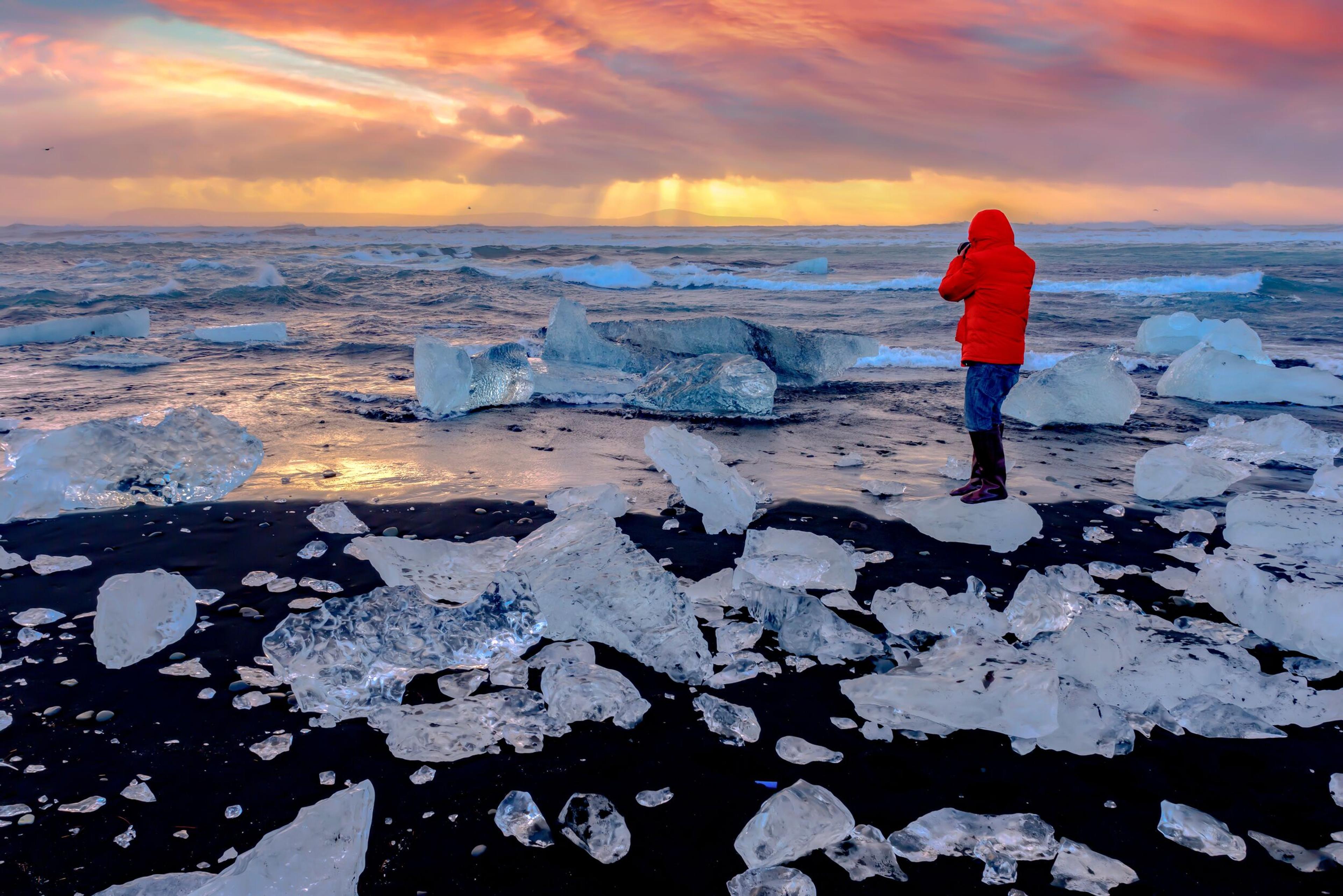 A person in a red jacket stands on a black sand beach scattered with ice chunks under a sunset sky.