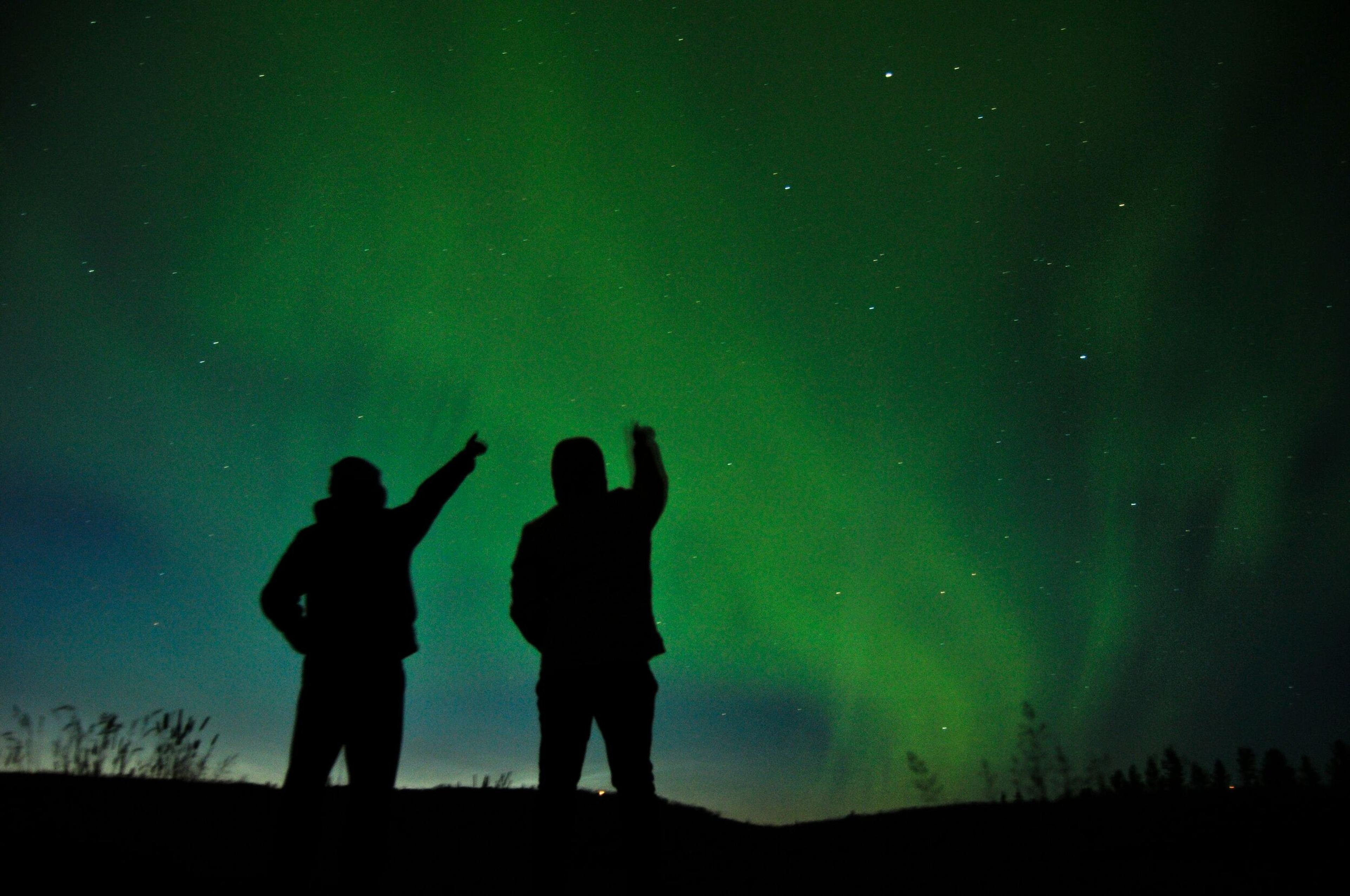  Silhouettes of two people standing beneath the green glow of the northern lights, pointing towards the sky.