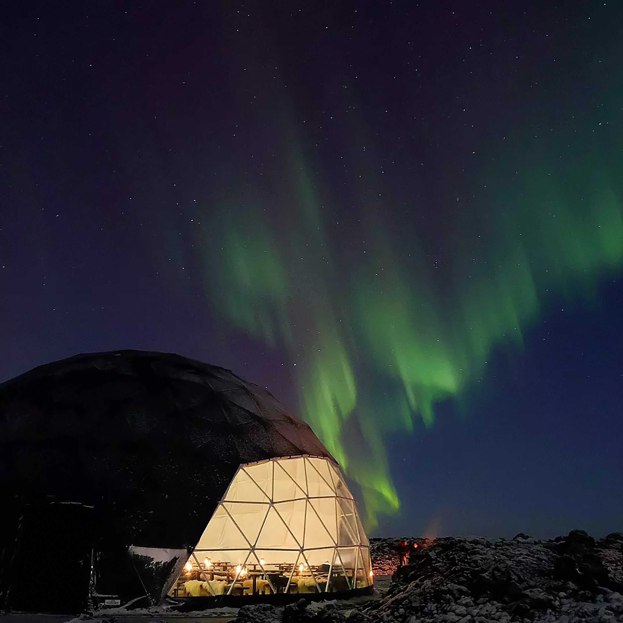 A geodesic dome at Aurora Basecamp is illuminated from within, nestled under a vivid display of green Northern Lights stretching across a dark, star-filled sky. 