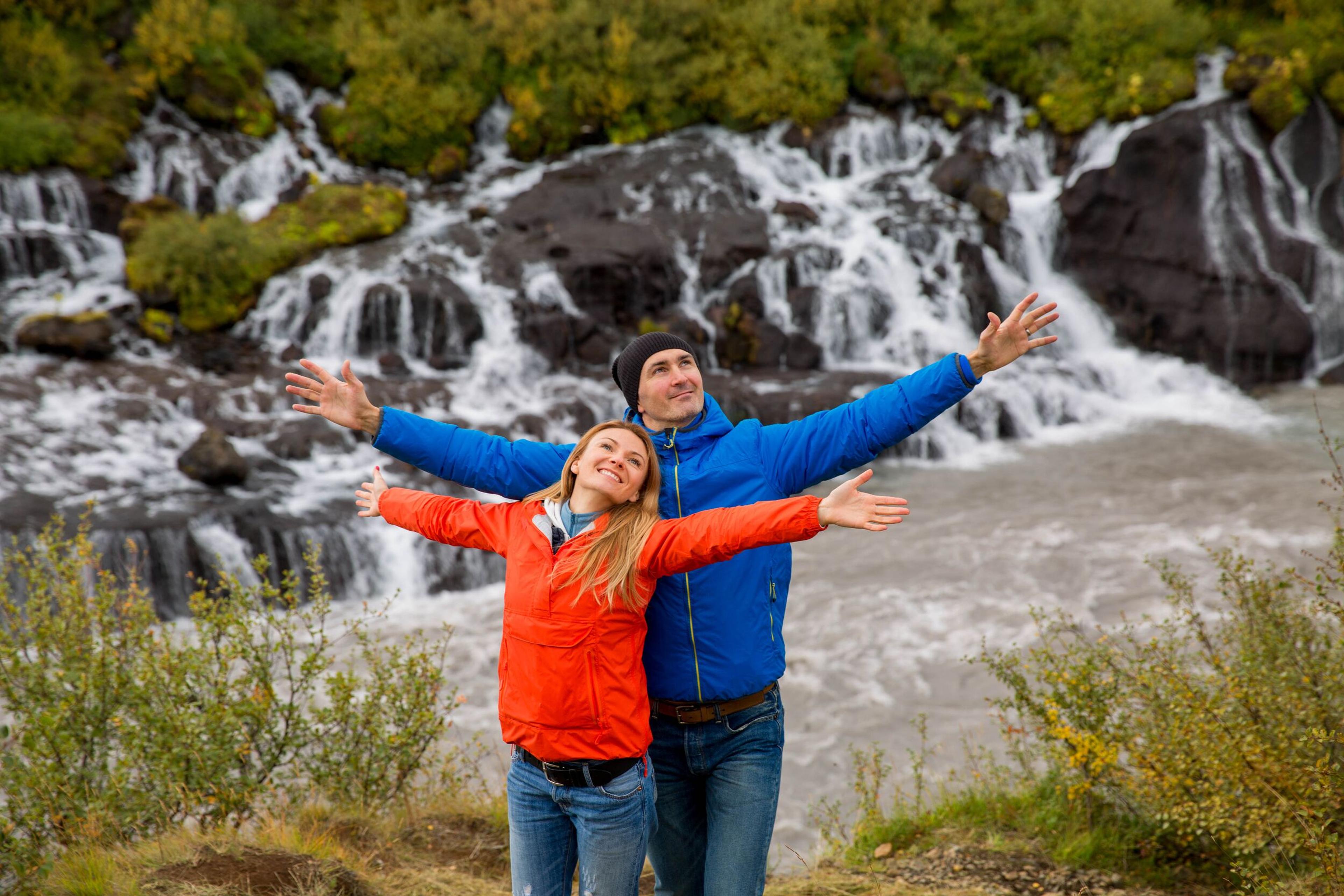 A joyful couple standing in front of Hraunfossar waterfalls, with arms outstretched, surrounded by lush greenery and cascading streams on Iceland's Silver Circle.