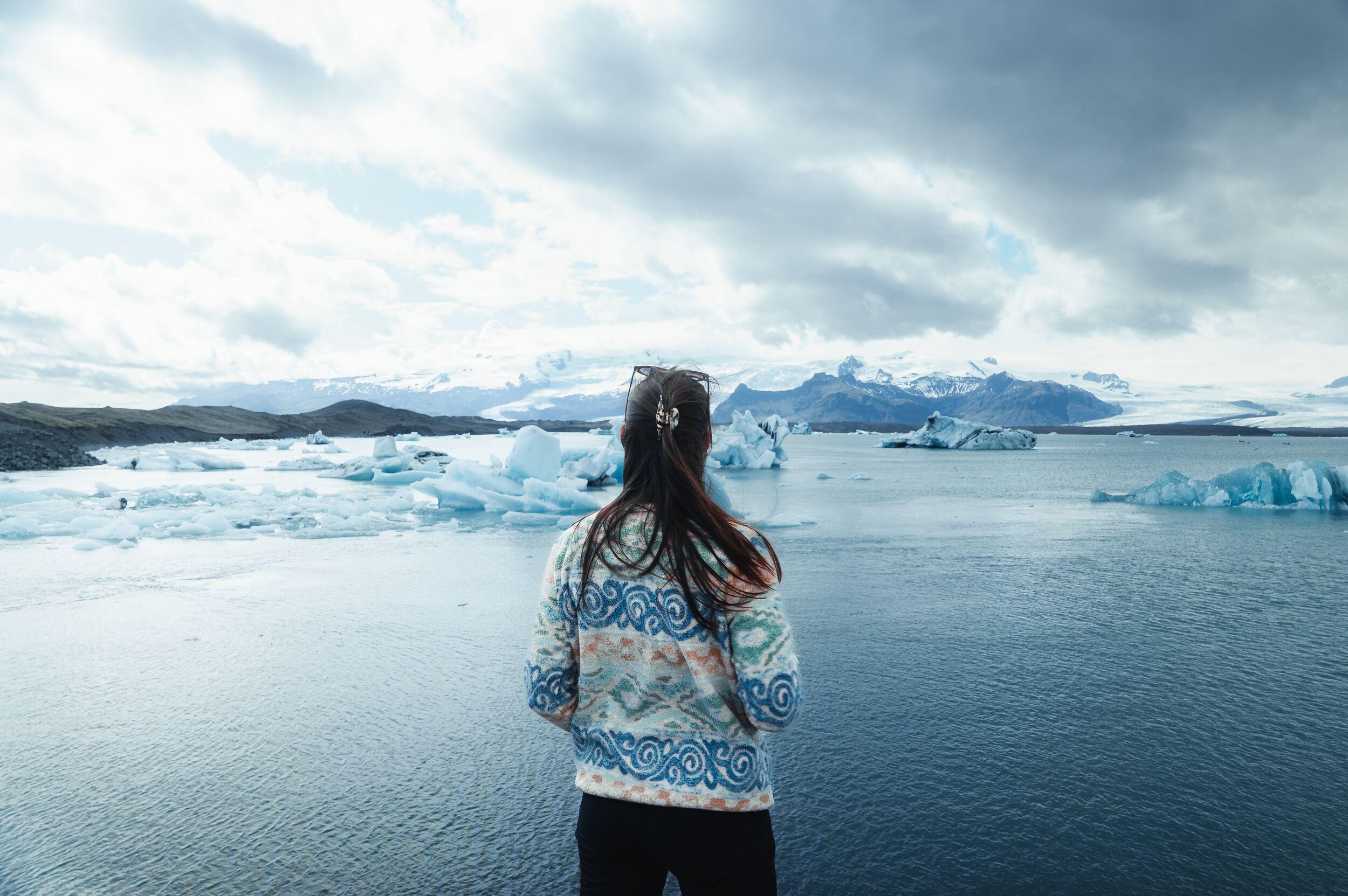 A person from behind looks out at an ice-filled glacial lagoon with mountains in the distance.