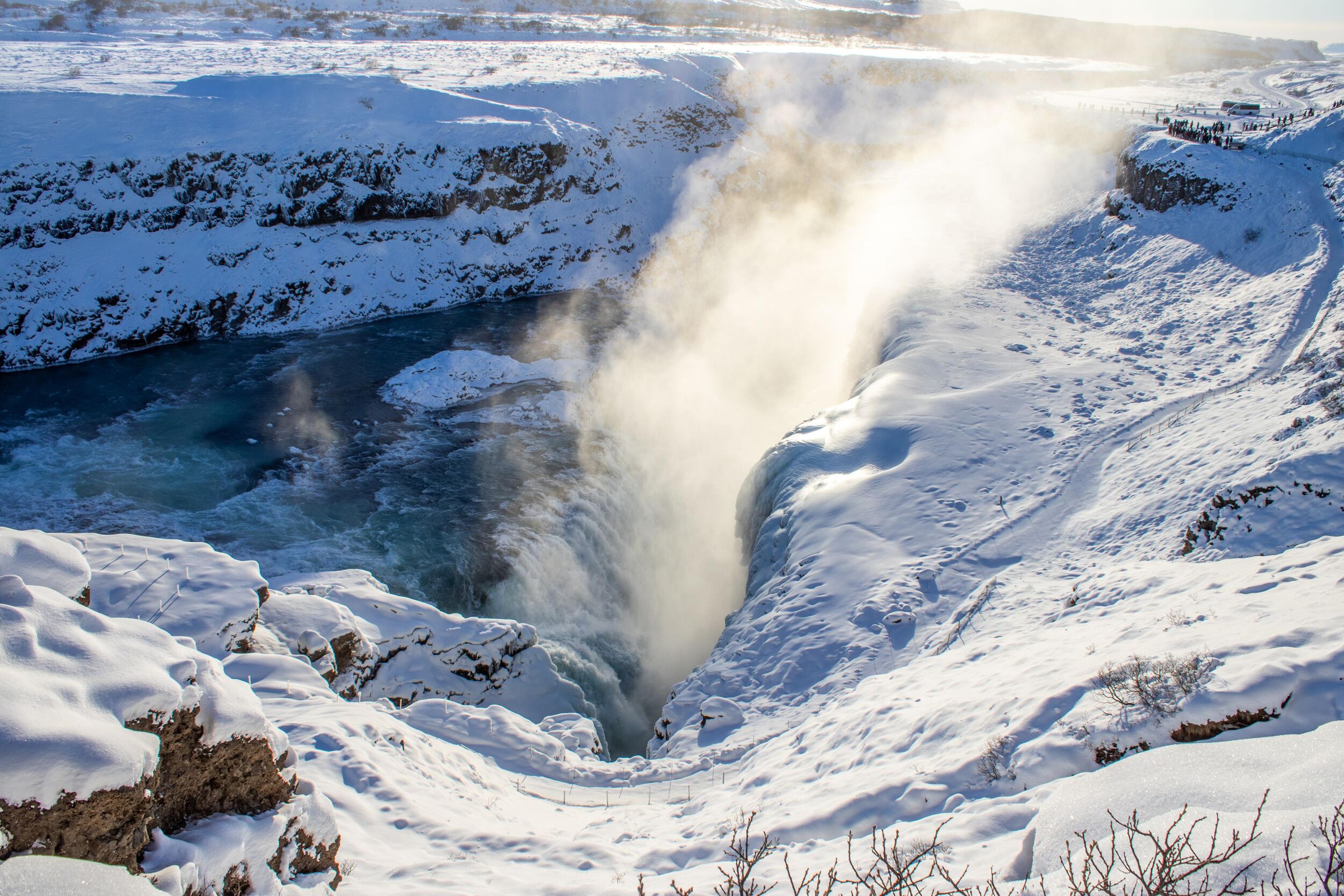 A powerful waterfall plunges into a snowy canyon, with mist rising from the churning blue water.