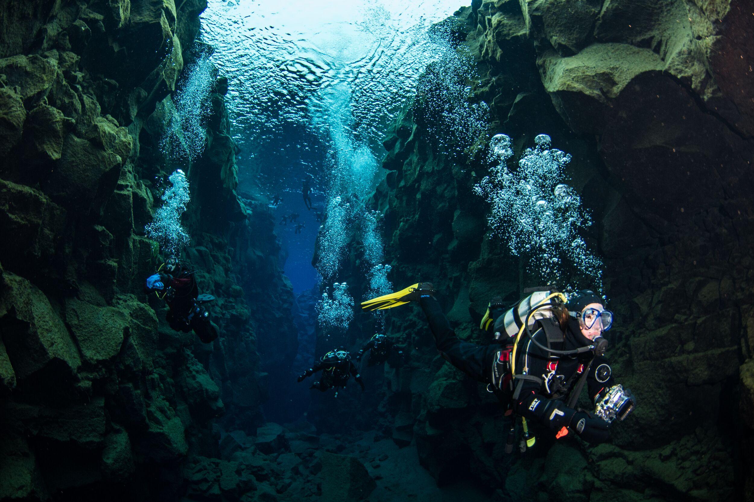 Scuba divers explore a narrow underwater canyon, with bubbles rising.