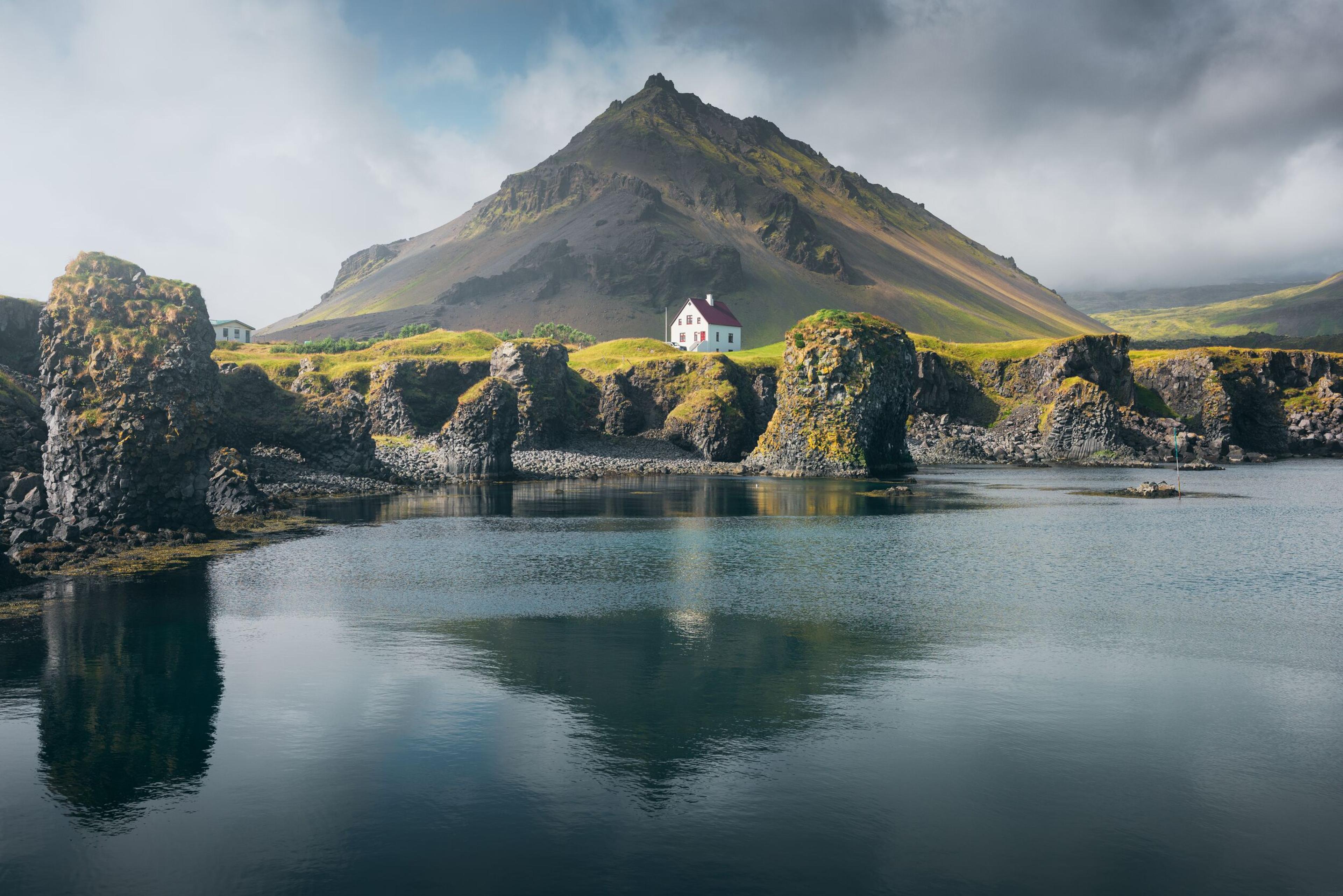A serene lake reflects jagged cliffs and a solitary white house with a red roof, nestled against a majestic mountain backdrop under a partially cloudy sky.