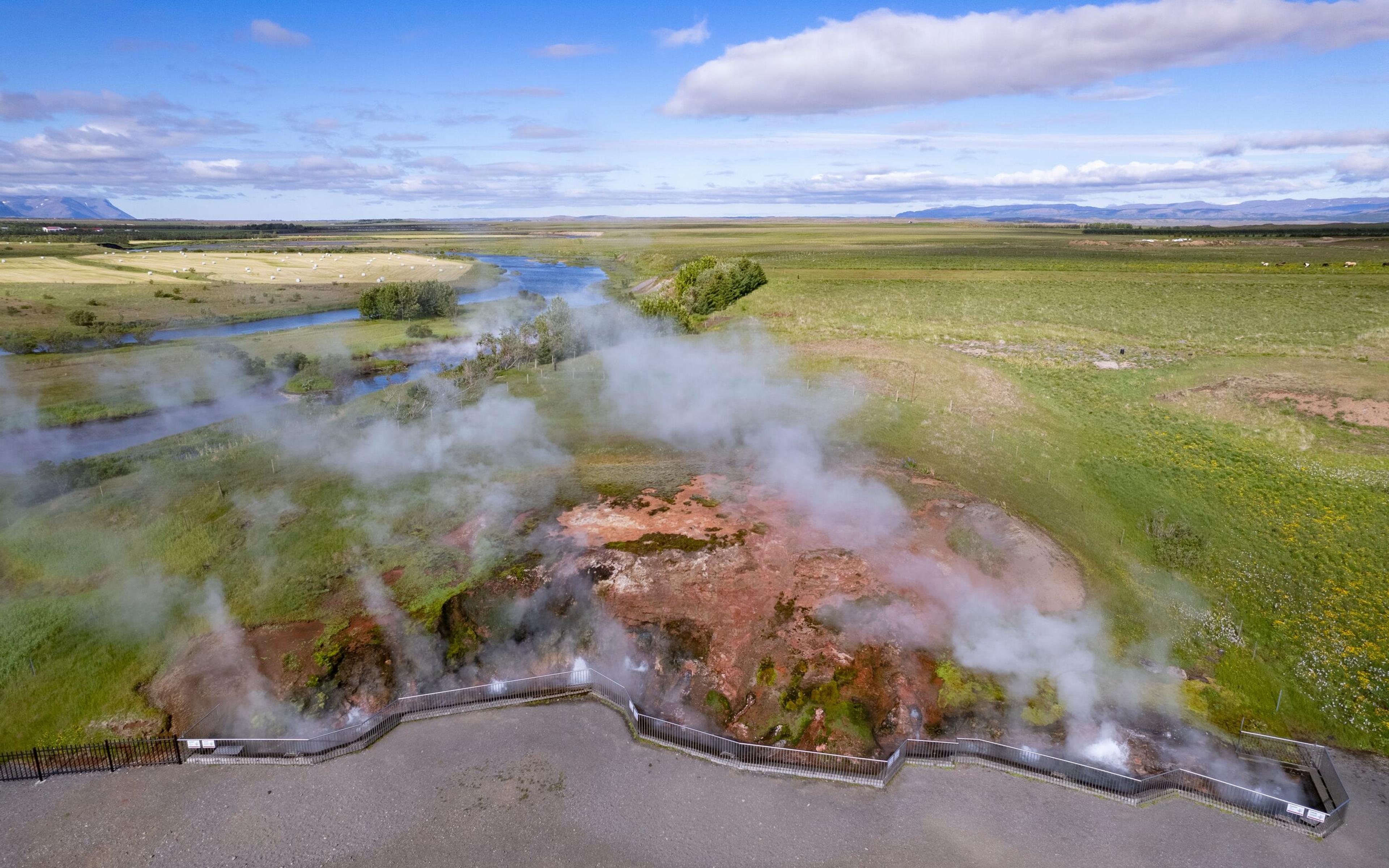 An aerial view of Deildartunguhver geothermal hot spring on the Silver Circle, featuring steaming vents surrounded by moss-covered rocks and vibrant geothermal colors, with visitors observing from a safe fenced platform.