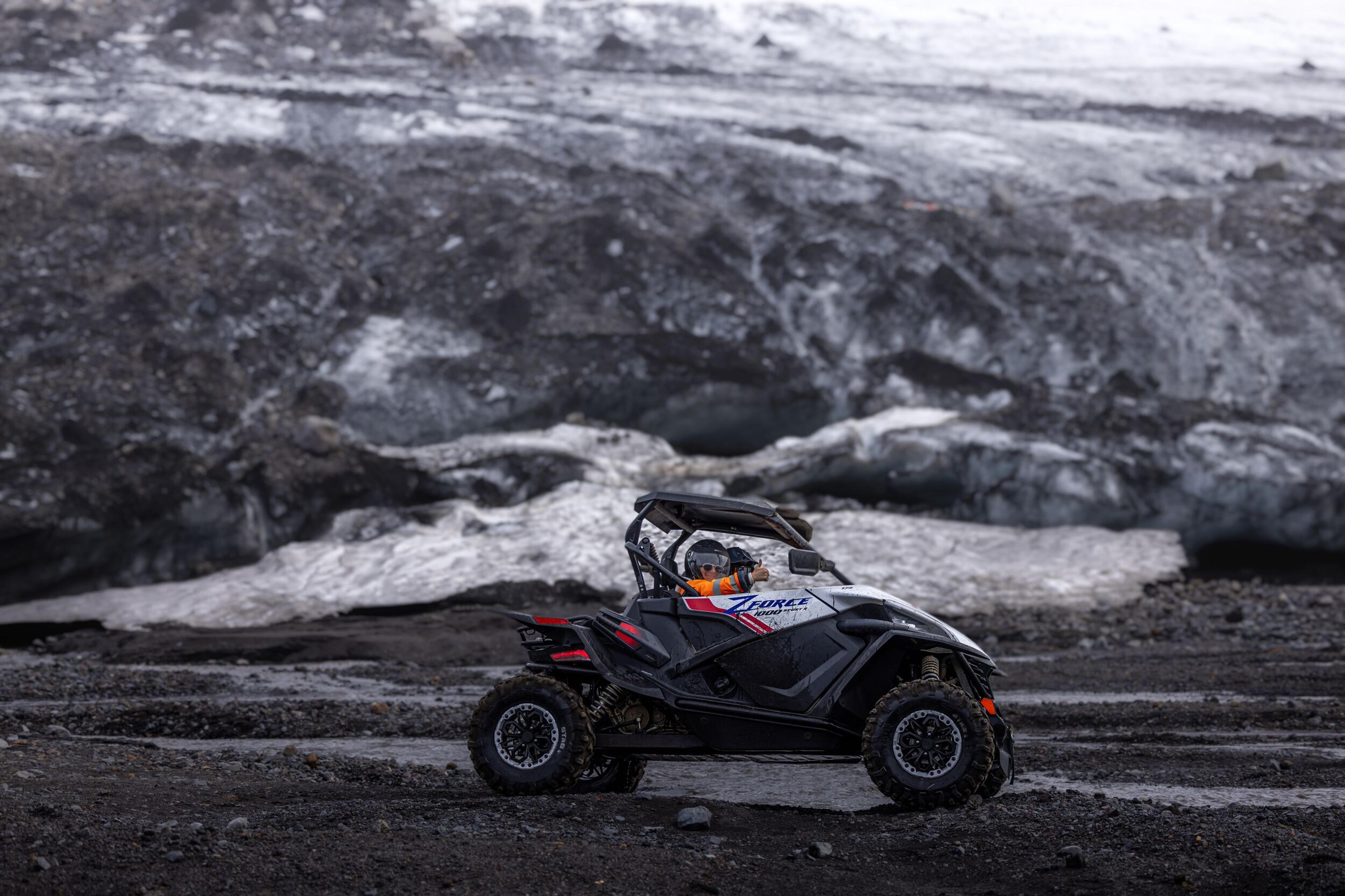 A black and orange off-road buggy is parked on dark volcanic terrain in front of a massive, icy glacier, blending adventure with Iceland’s extreme landscapes.