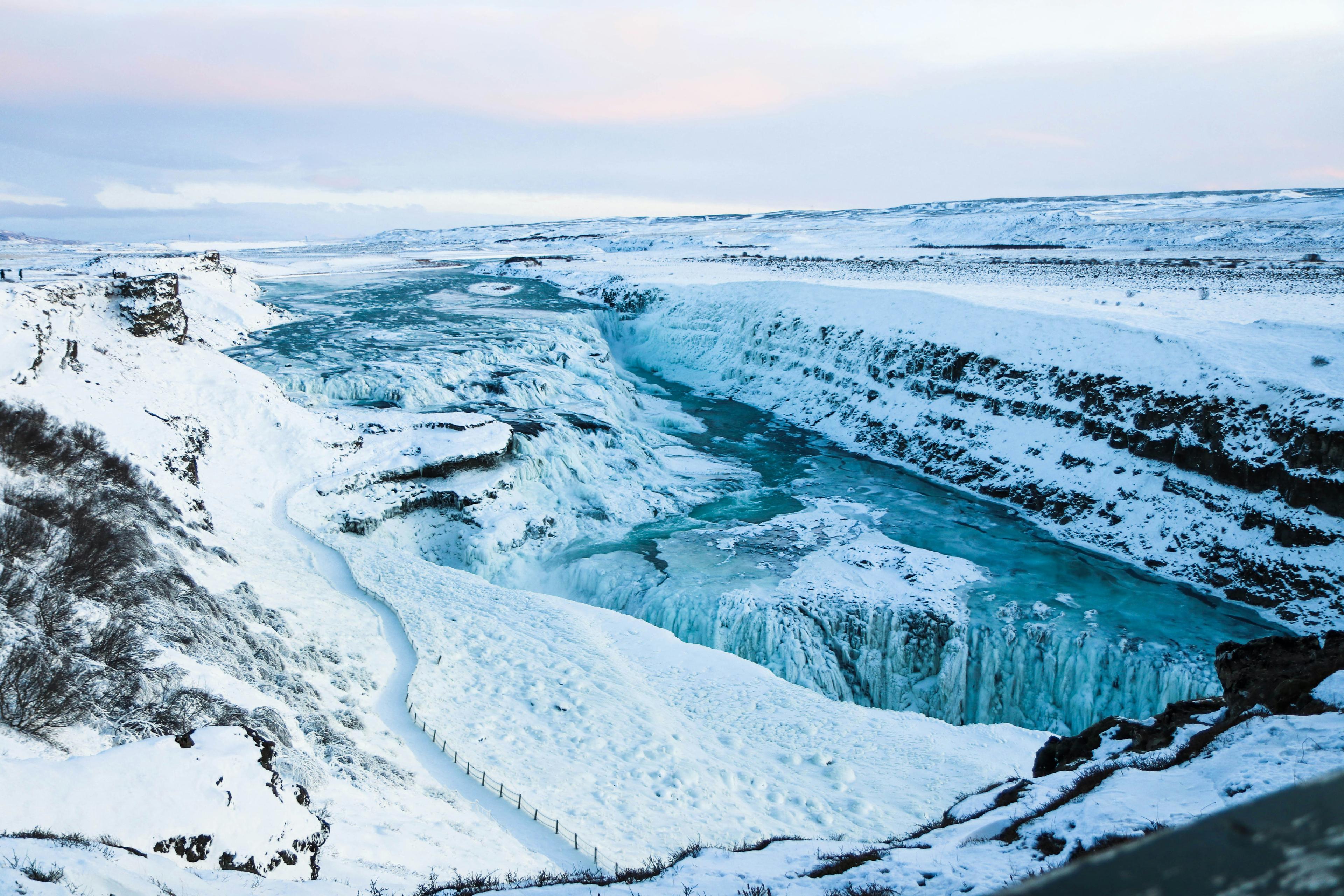  A stunning winter scene of Gullfoss waterfall in Iceland, partially frozen over. The powerful cascades are now solid ice, forming intricate patterns and icicles hanging from the edges. The rugged canyon is blanketed in snow, with the icy river below reflecting the pale winter light. 