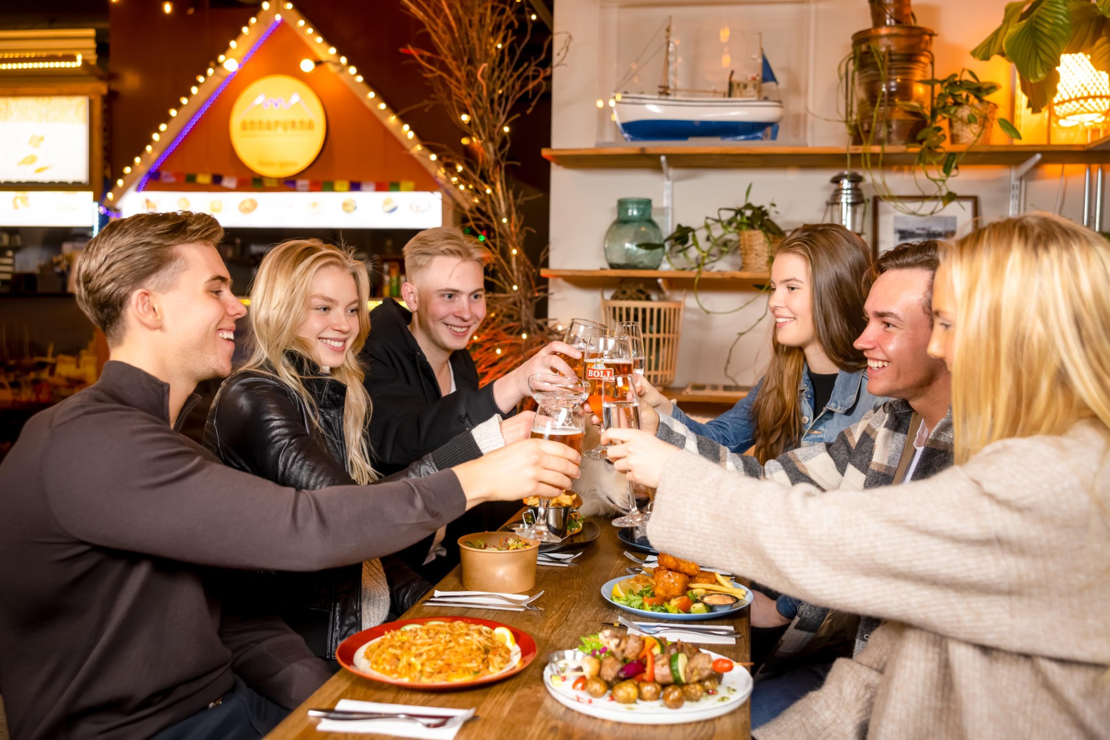 Friends toasting drinks at a Food Hall, enjoying a meal together with a variety of dishes on the table and festive decor in the background.