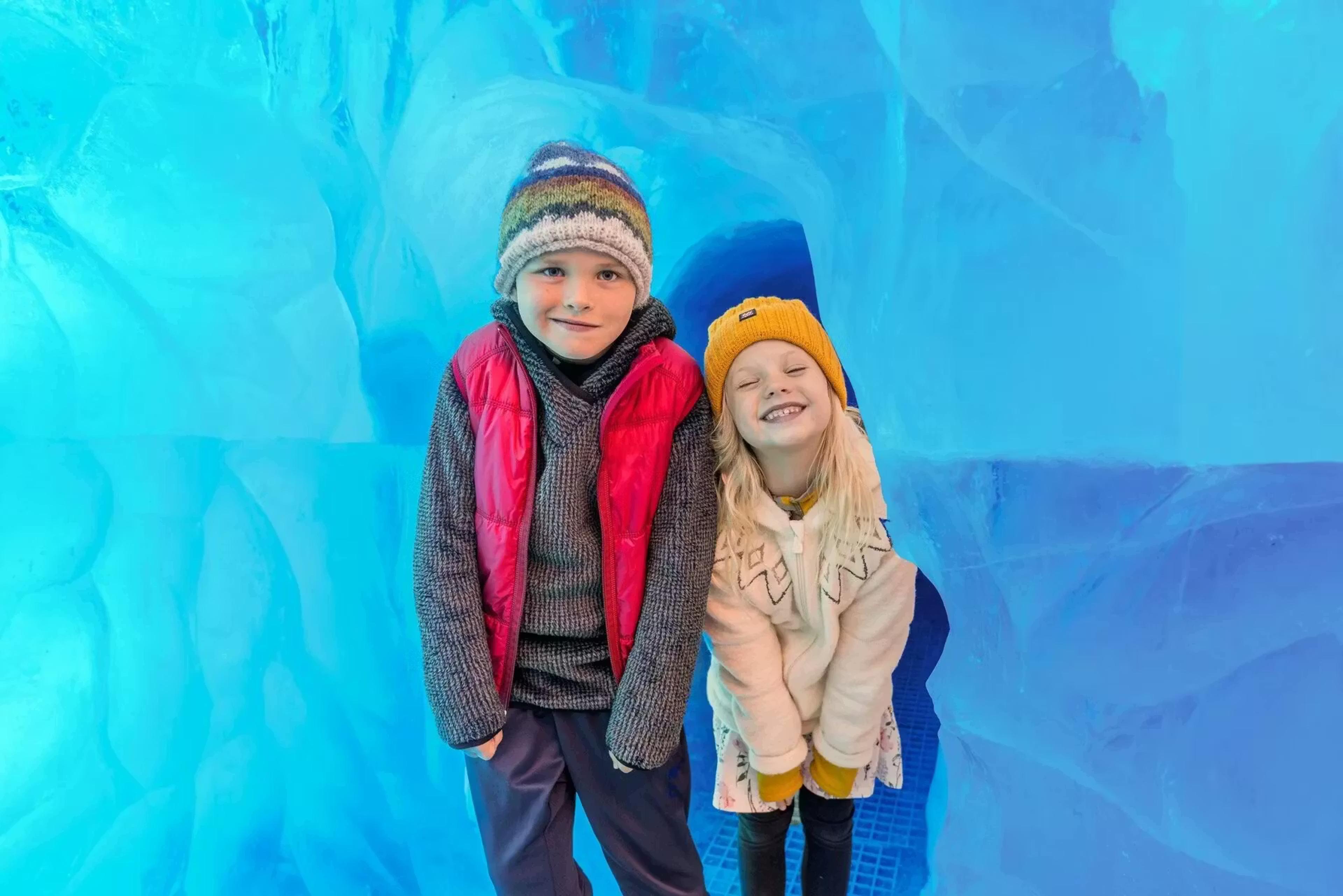 Two smiling children, a boy and a girl, stand against a vivid blue ice background.