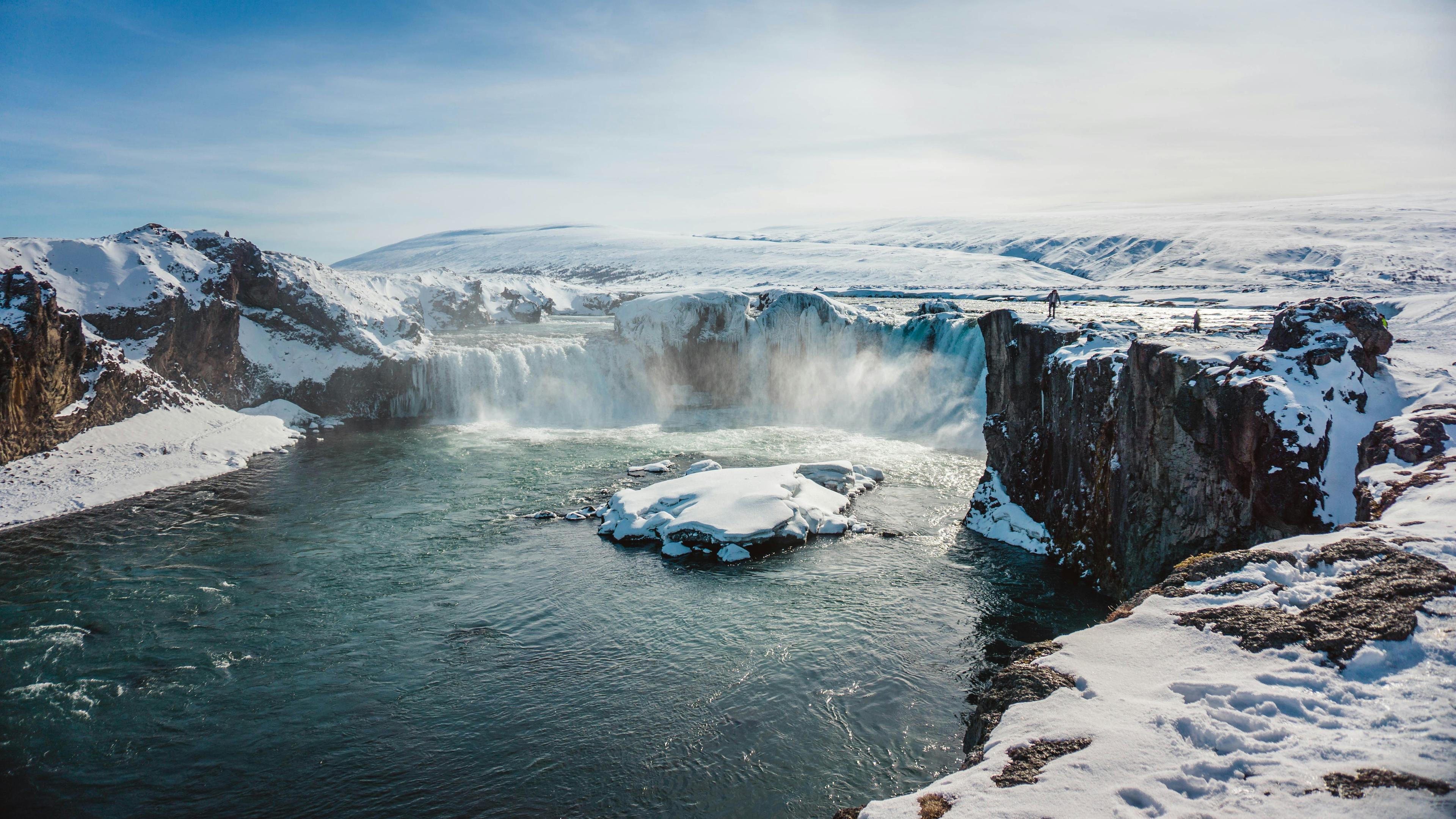 A wide waterfall flows into a river surrounded by snow-covered cliffs and rocks. 