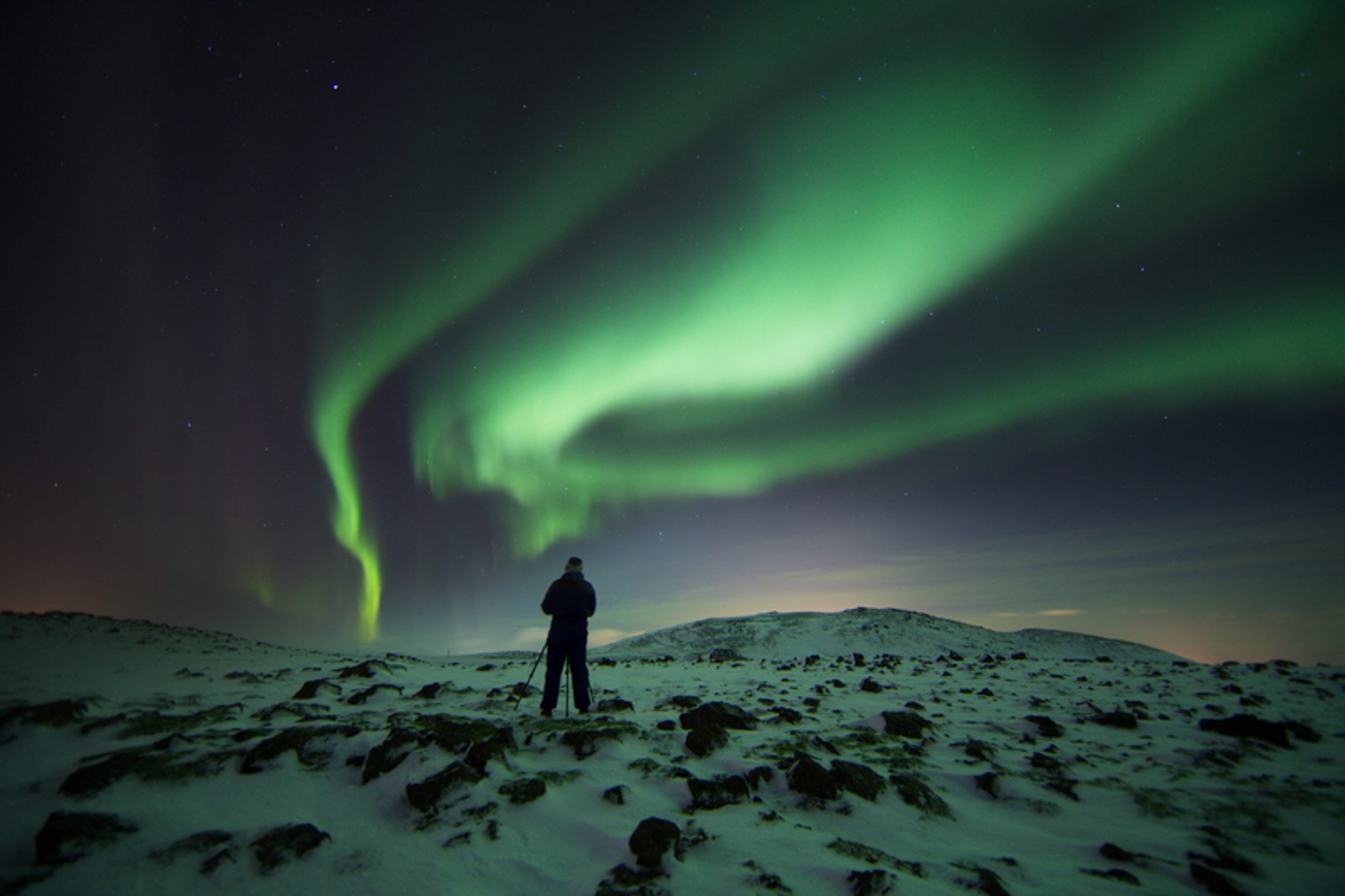  A lone figure stands on a snowy hill, gazing up at the vibrant green aurora borealis swirling in the night sky.