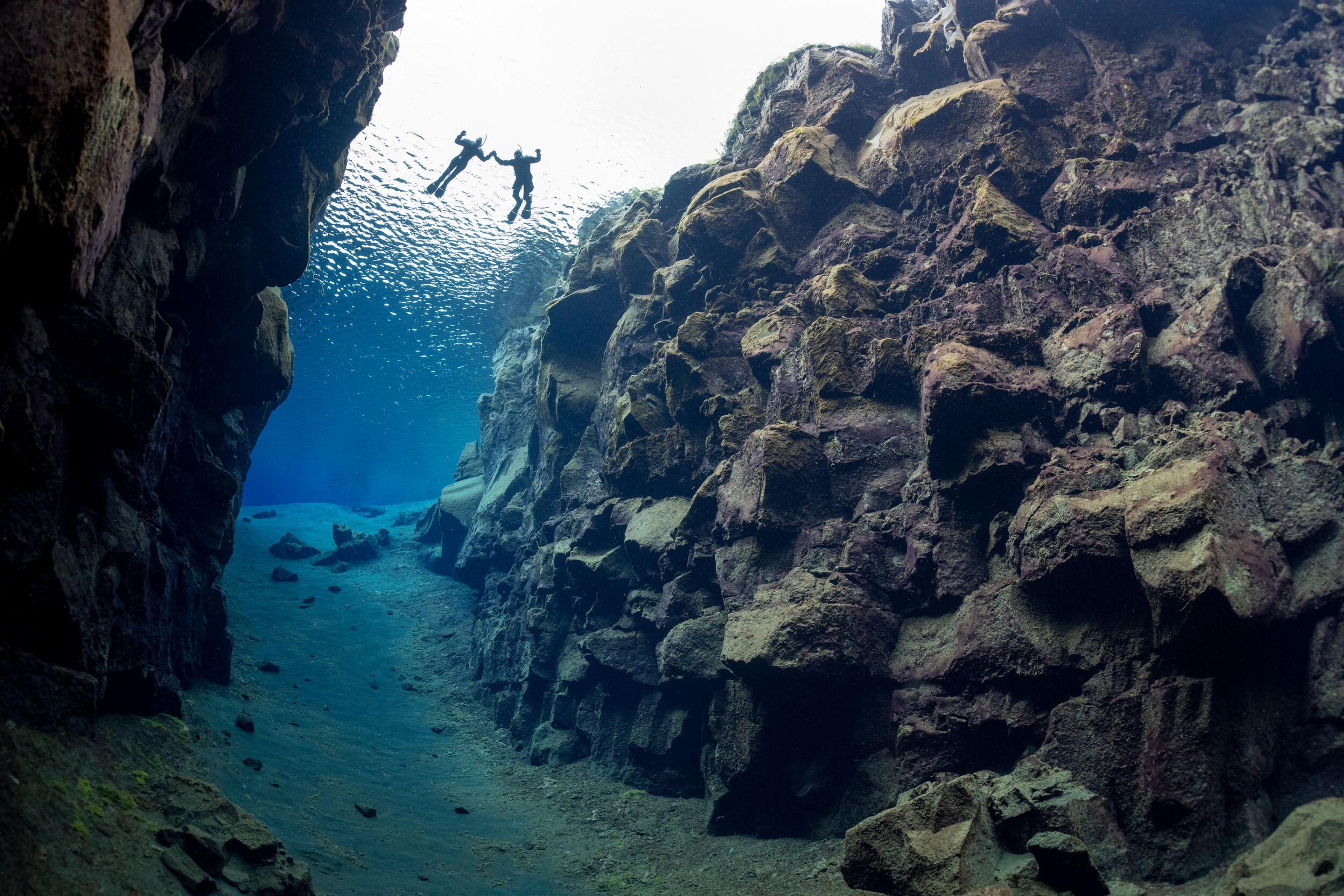 Two divers swim between the massive underwater rock formations of Silfra, with crystal-clear water and sunlight filtering from above.