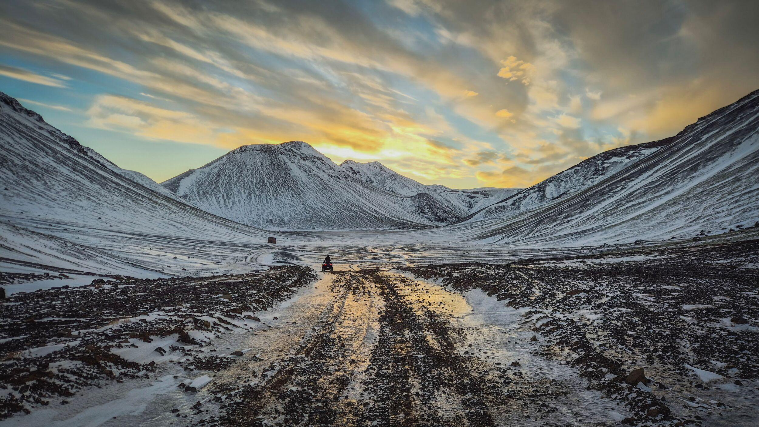 A person on a vehicle drives along a muddy, snowy road through a snow-dusted mountain valley under a golden sunset.