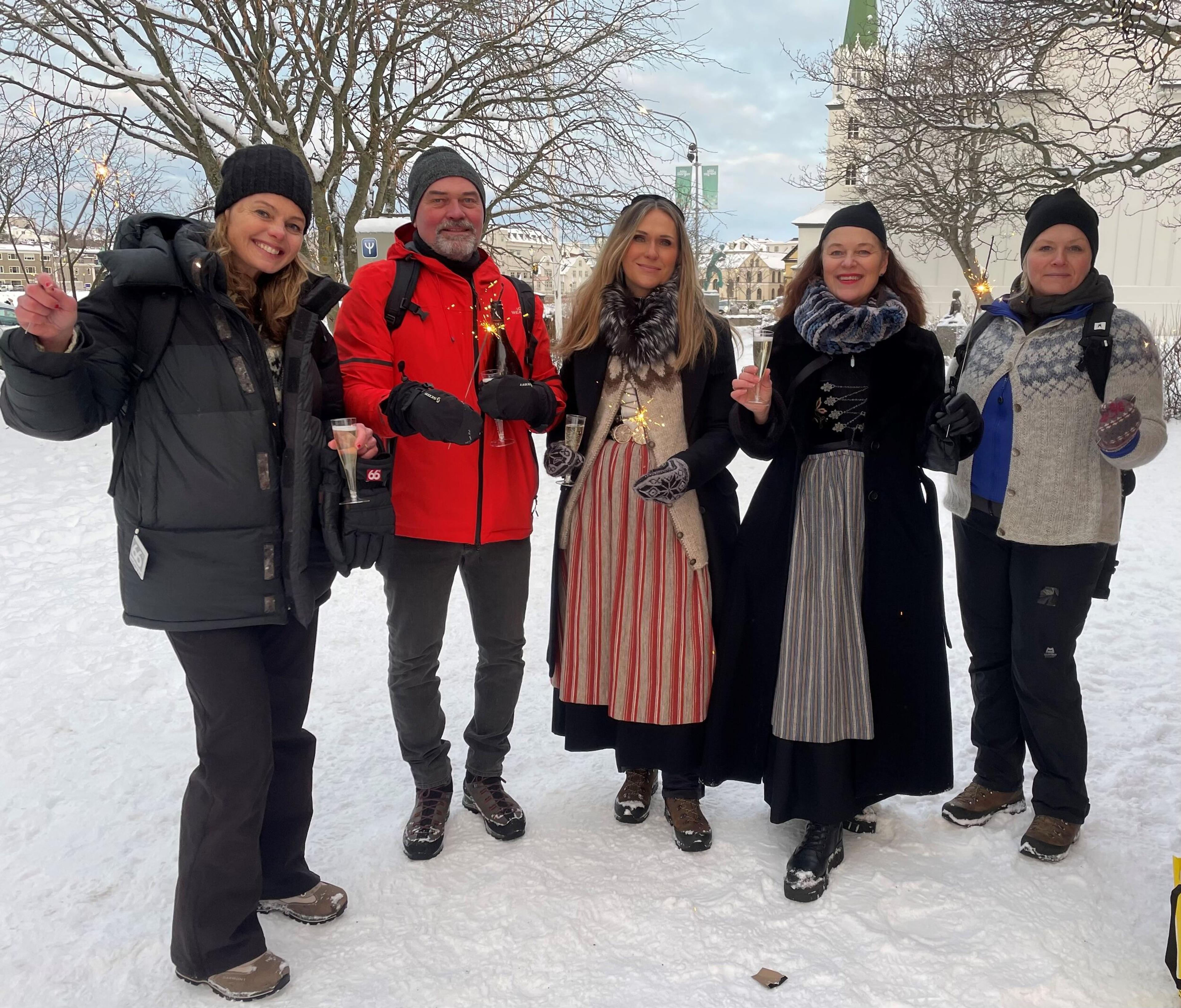 Five people, some in traditional dress, celebrating with sparklers and champagne in a snowy town.