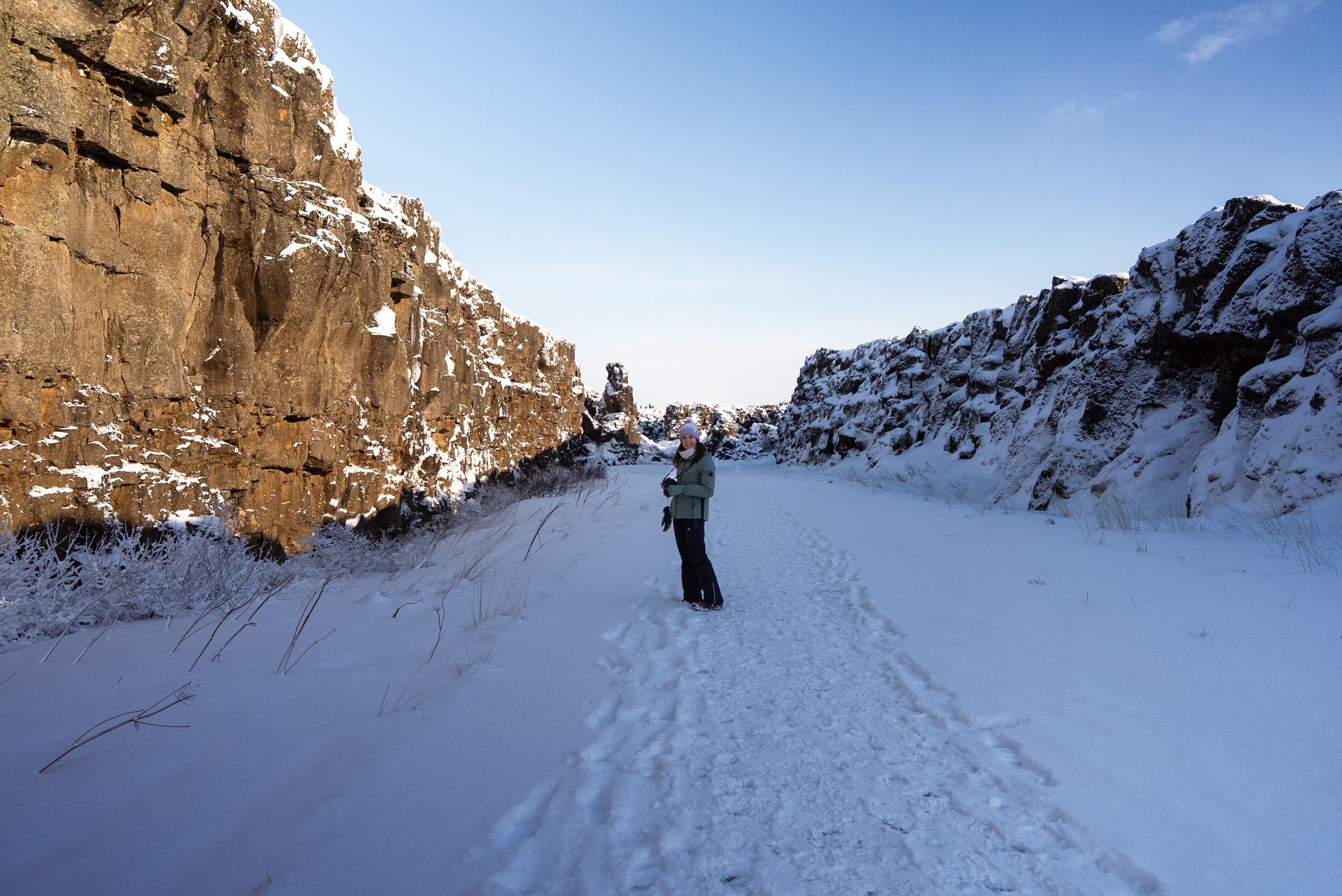 A person stands on a snowy path between rugged, snow-dusted cliffs under a clear blue sky.