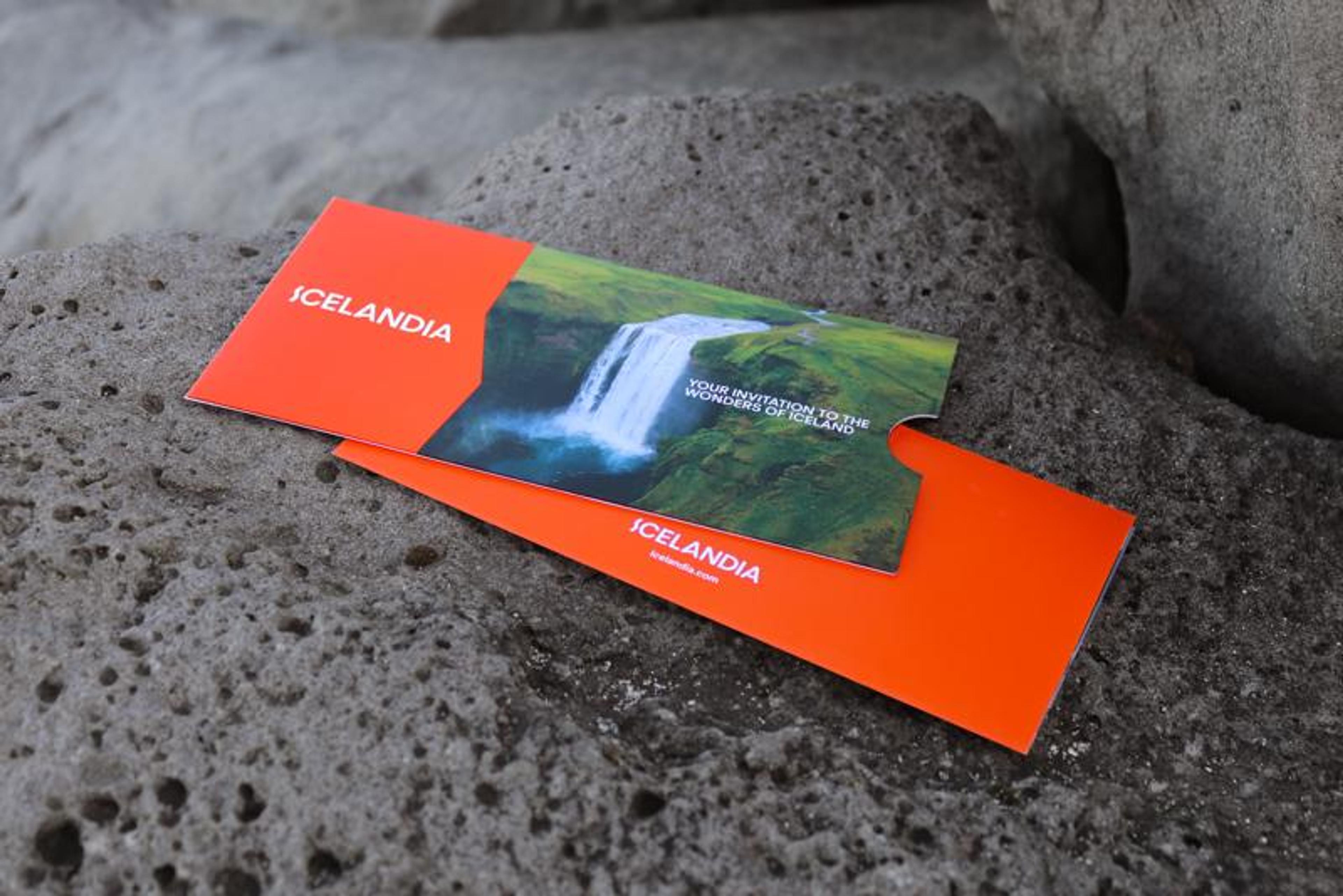 An orange ICELANDIA brochure featuring a waterfall image and travel invitation text, resting on a gray rock.