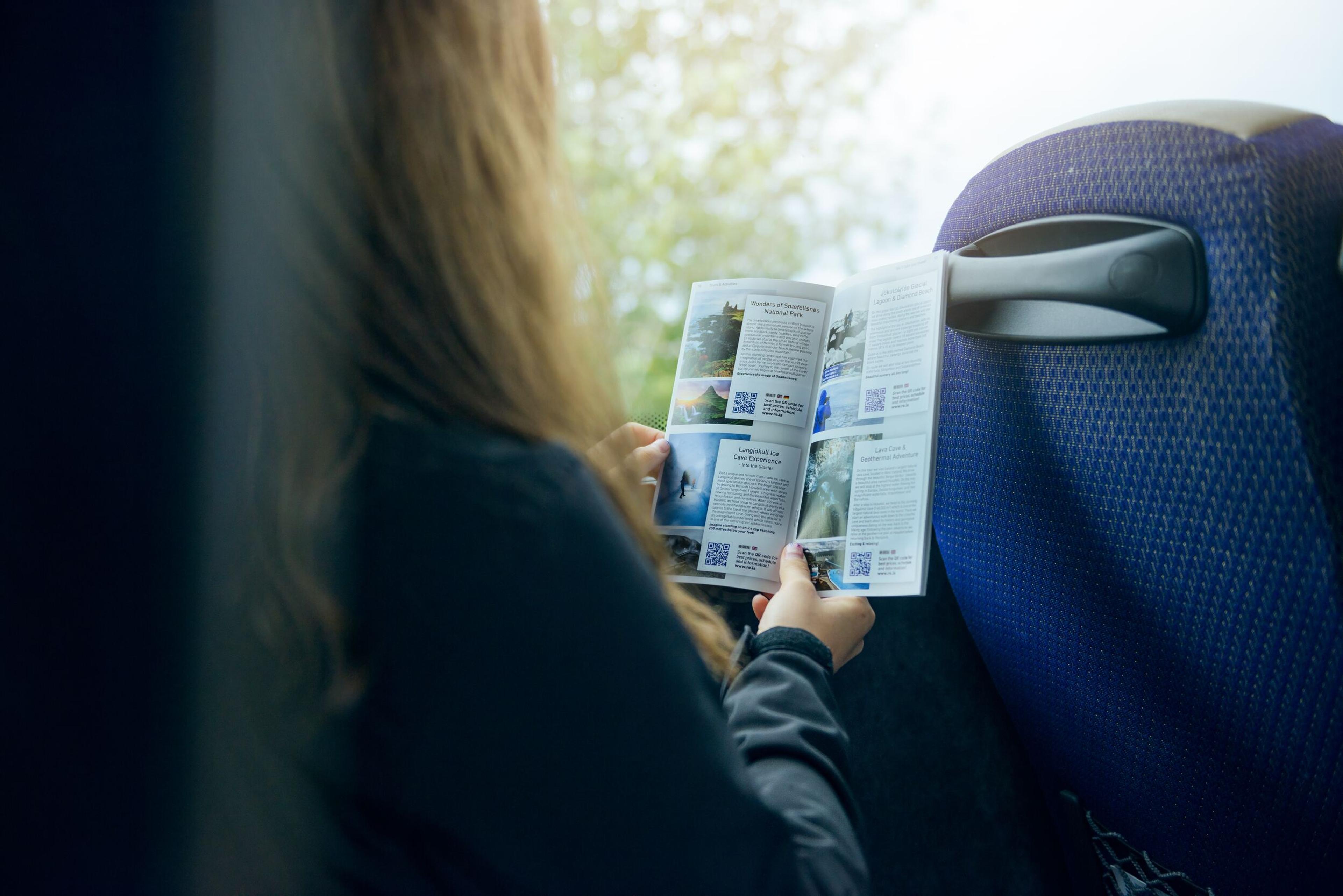 A person seated on a bus, reading a pamphlet or brochure, with a focus on the back of their head and the brochure.