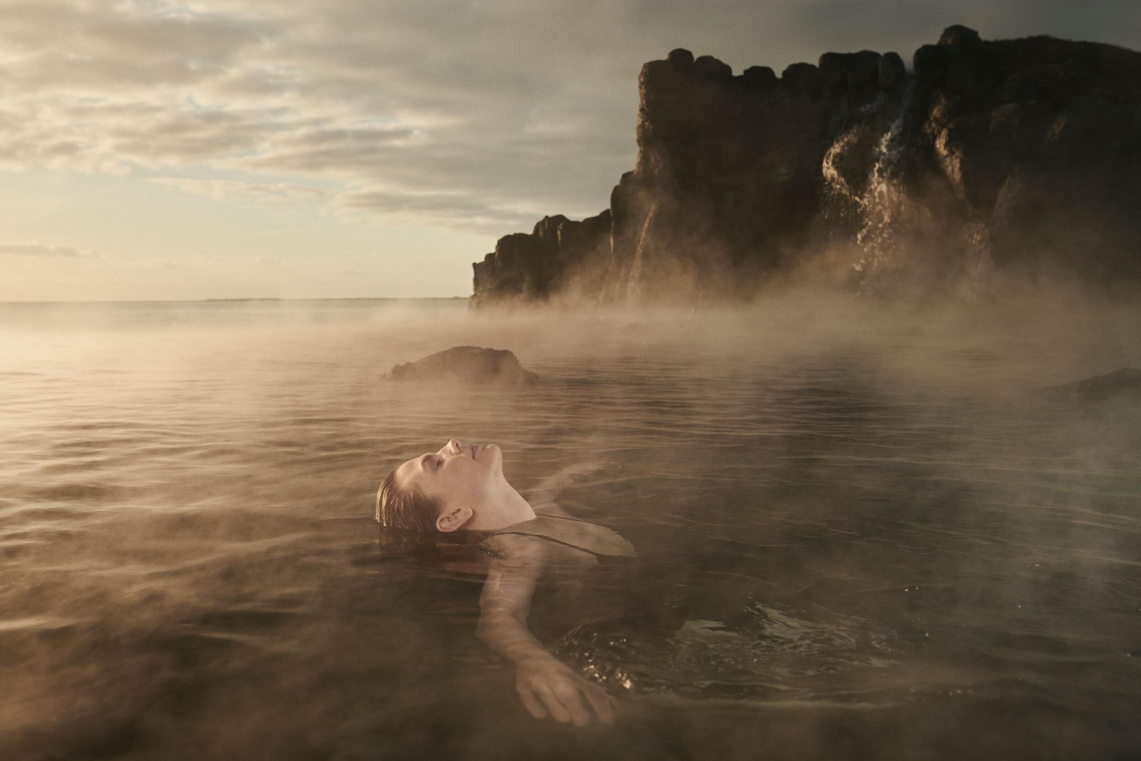 A woman serenely floating in warm waters, framed by towering cliffs, bathed in the golden hues of sunset.