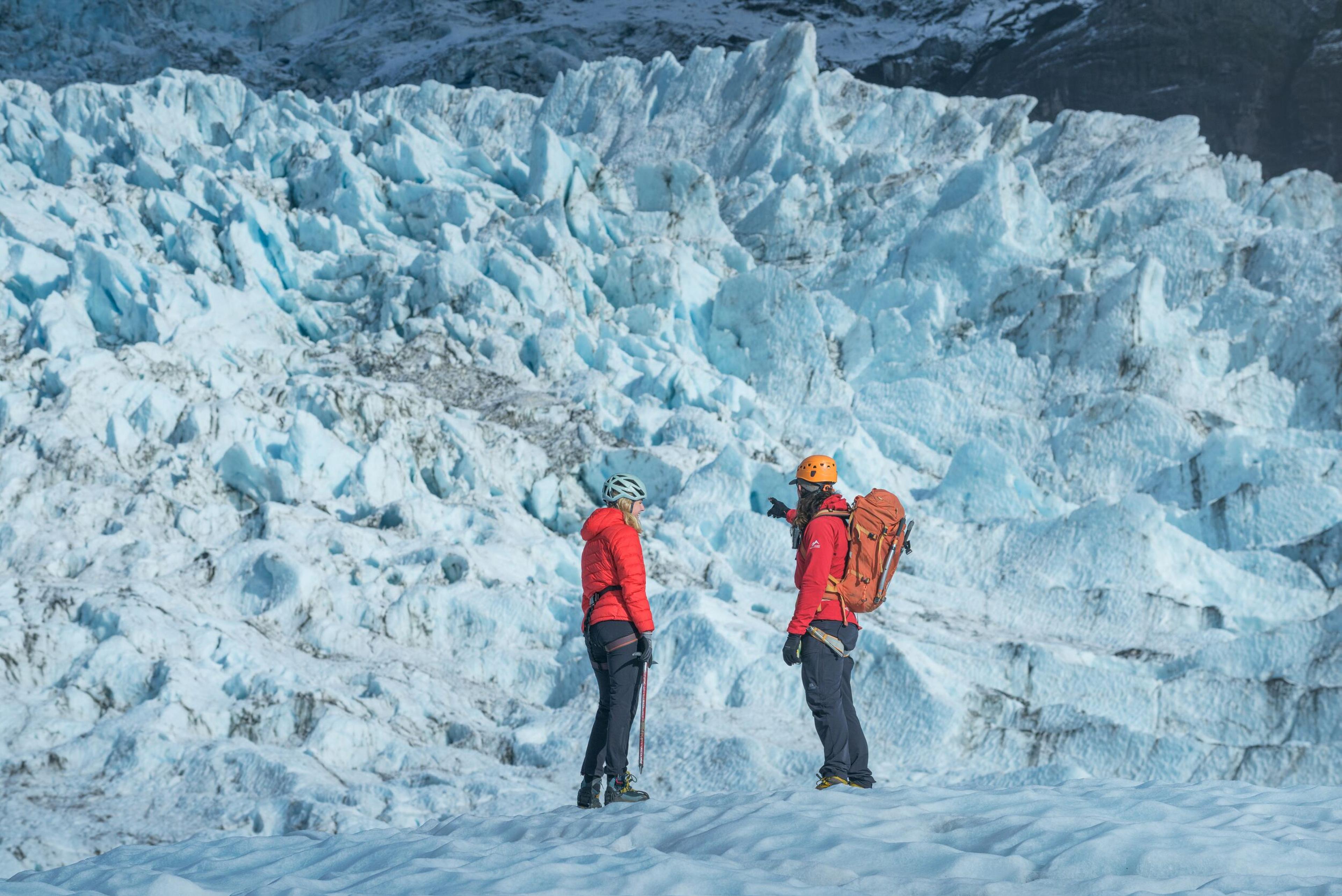 Two adventurers in vibrant red jackets and safety helmets stand atop a brilliant blue glacier, with jagged ice formations providing a dramatic backdrop