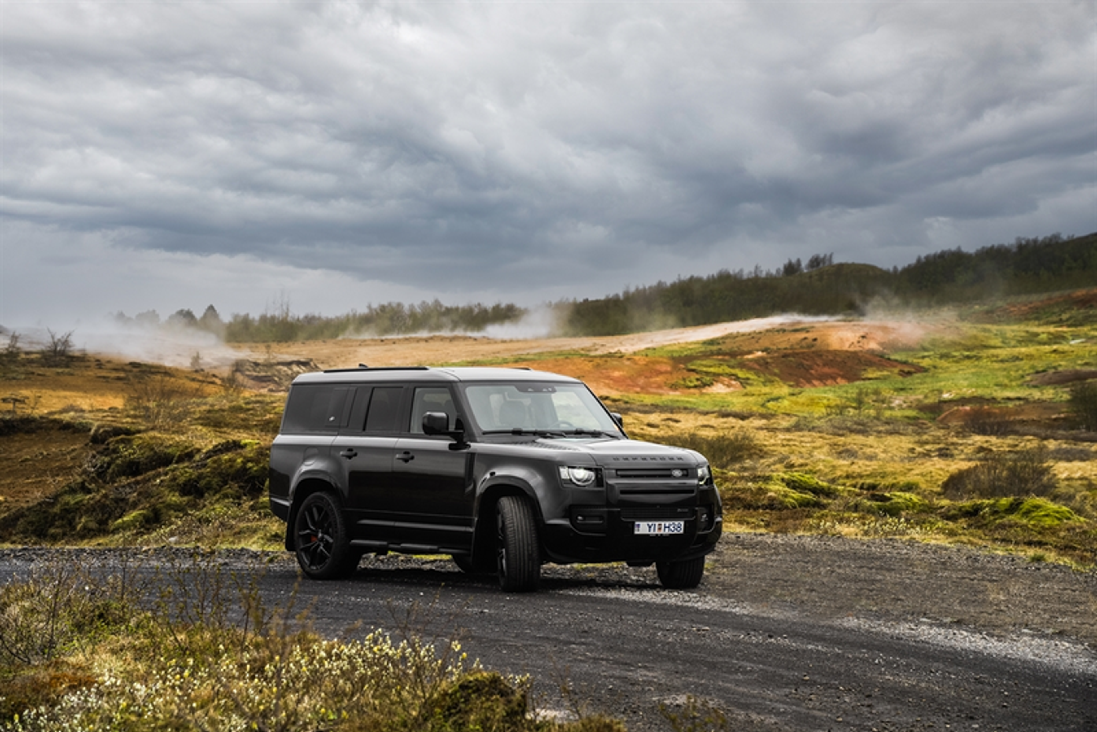 : A sleek black SUV on a rugged gravel road, surrounded by geothermal steam and vibrant Icelandic terrain under an overcast sky. Ideal for exploring the Golden Circle on a private tour.