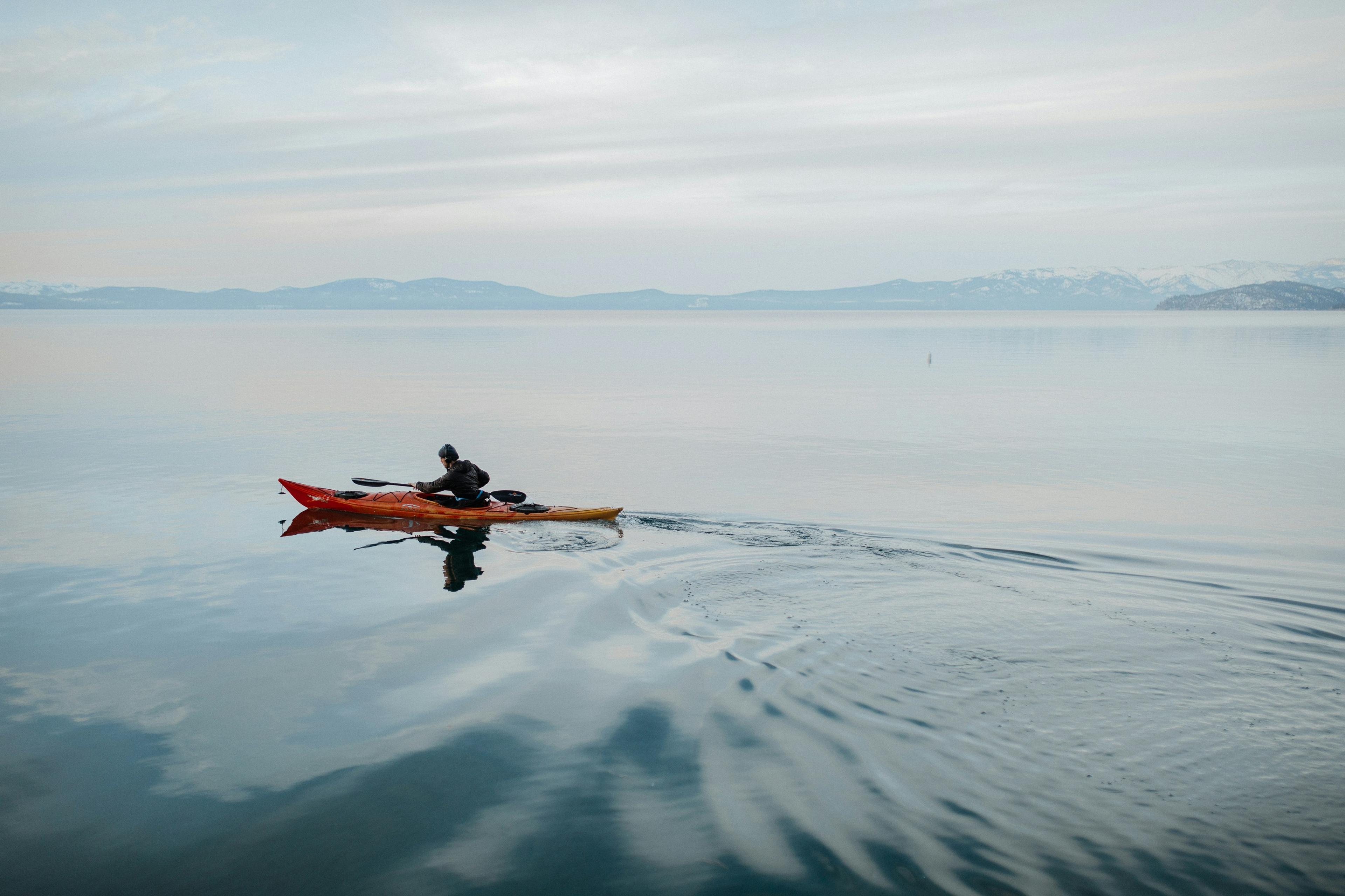 A lone kayaker in a red and yellow kayak paddles on serene waters with distant mountains under a cloudy sky, highlighting kayaking in Iceland.