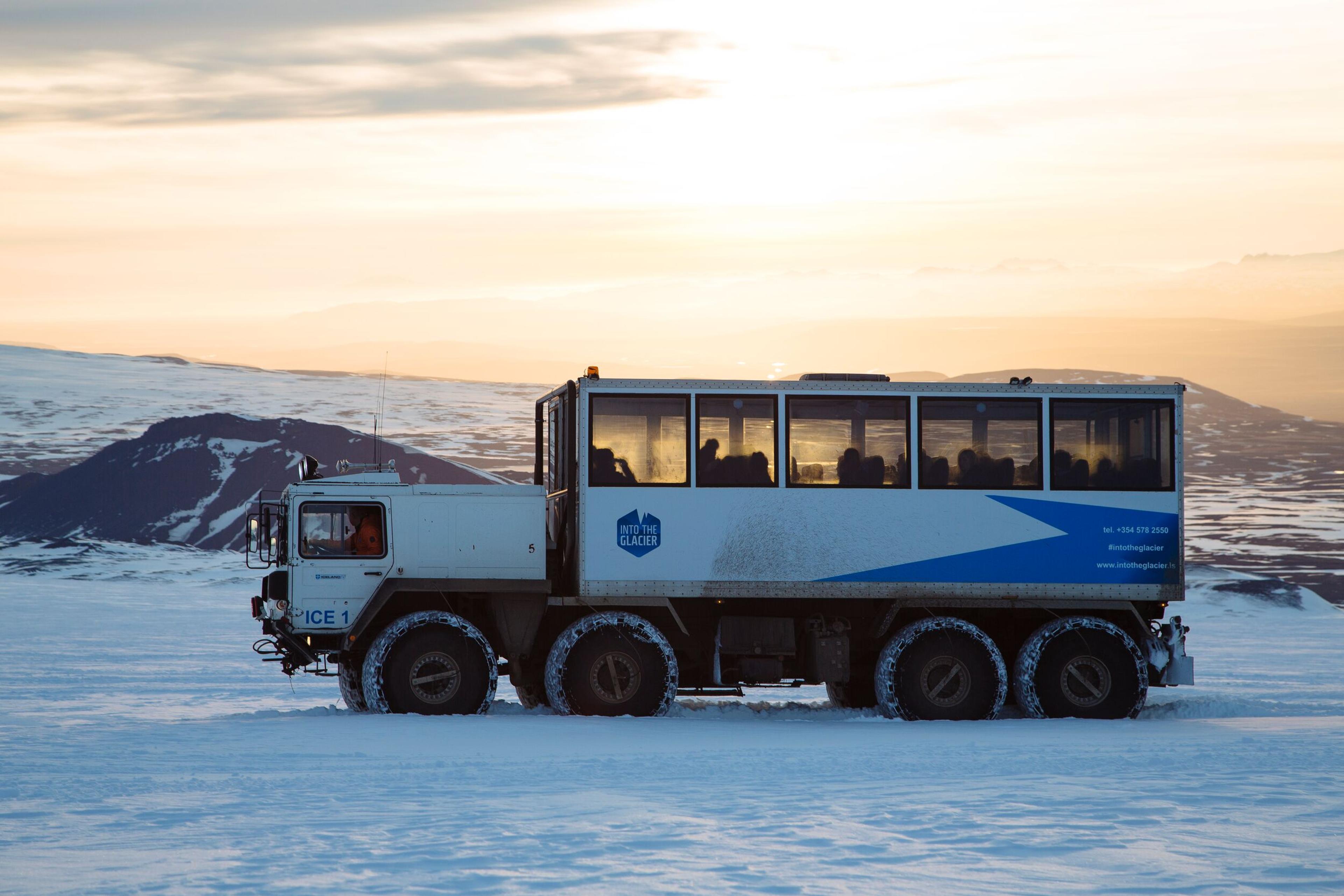 A massive off-road monster truck parked on the snowy expanse of Langjökull glacier, set against a backdrop of rugged Icelandic terrain and a pastel sky.