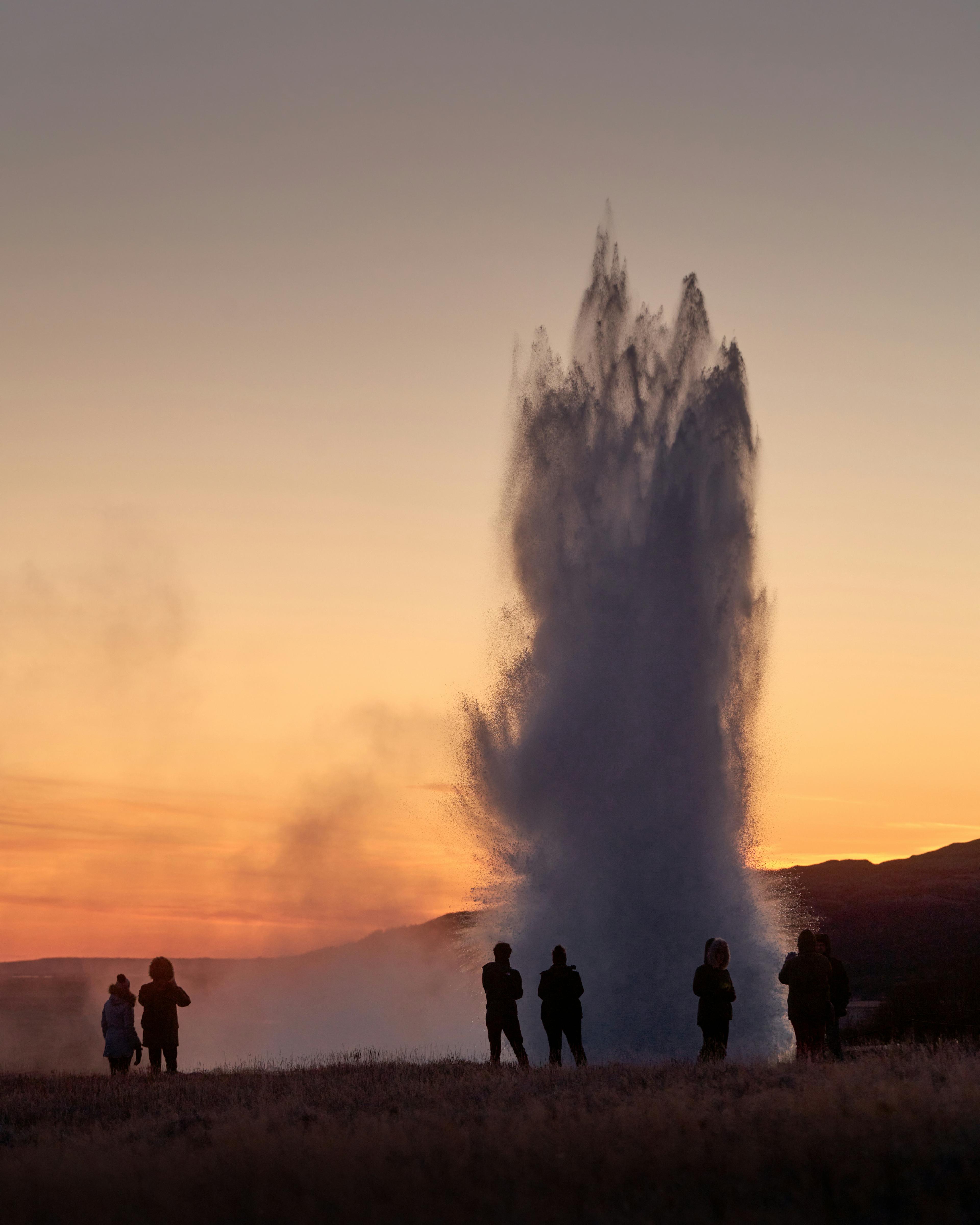 Silhouetted figures stand in awe as a geyser erupts into the twilight sky, its plume highlighted by the warm hues of sunset, a captivating moment of natural wonder.