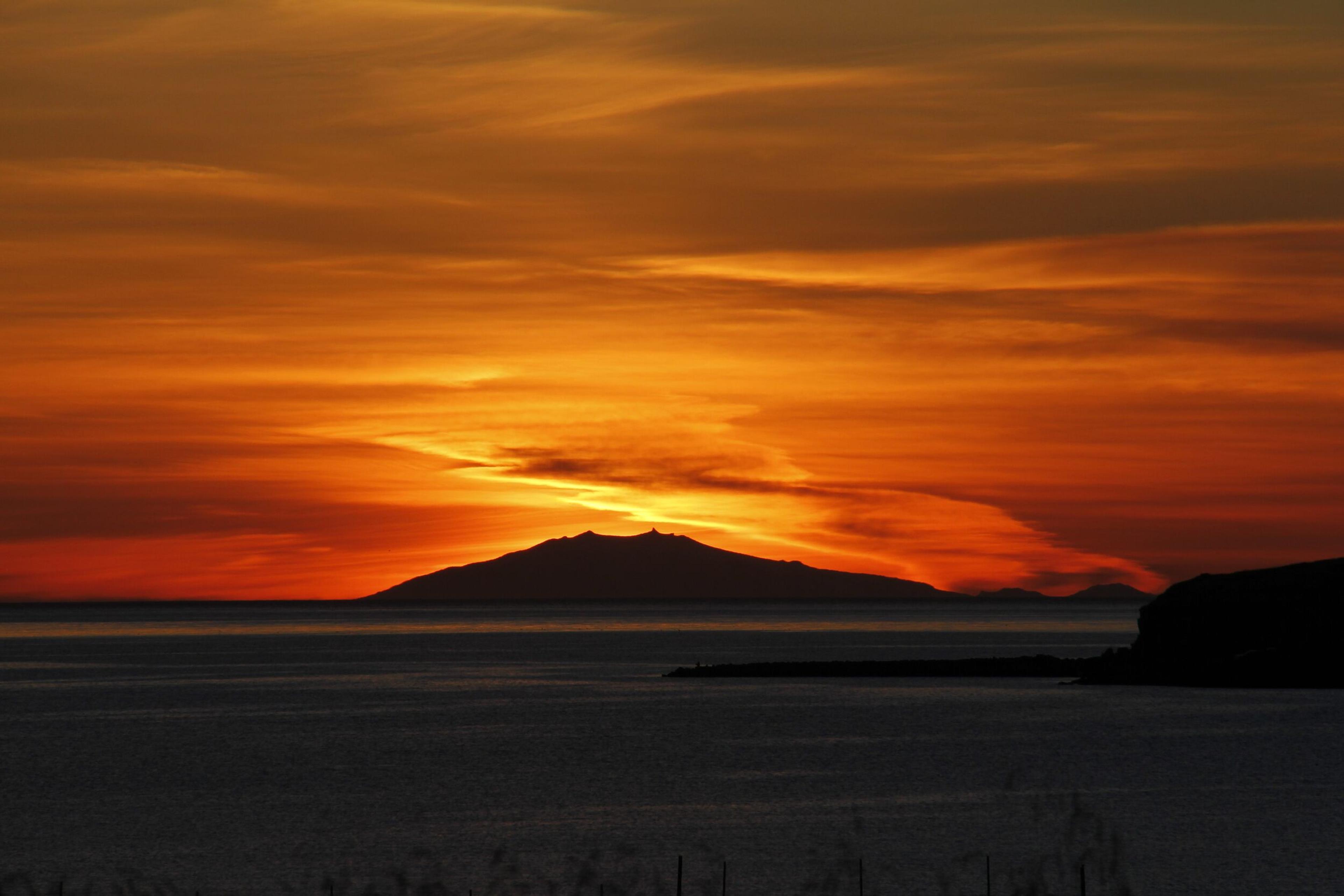 A breathtaking sunset paints the sky in fiery orange hues above a silhouetted mountain range, casting a dramatic backdrop over the calm sea.