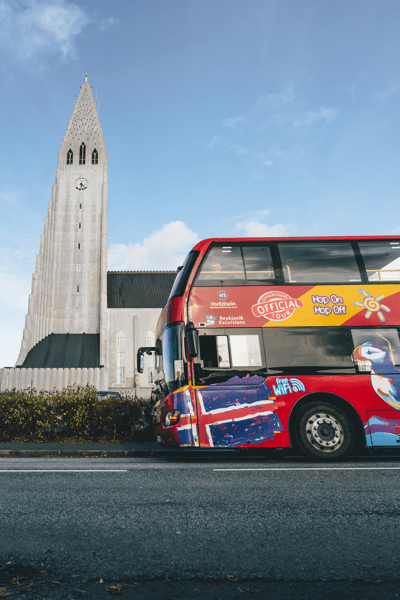 A red double-decker "Hop On Hop Off" bus with an Icelandic flag design sits on a street in front of Hallgrímskirkja church under a blue sky.