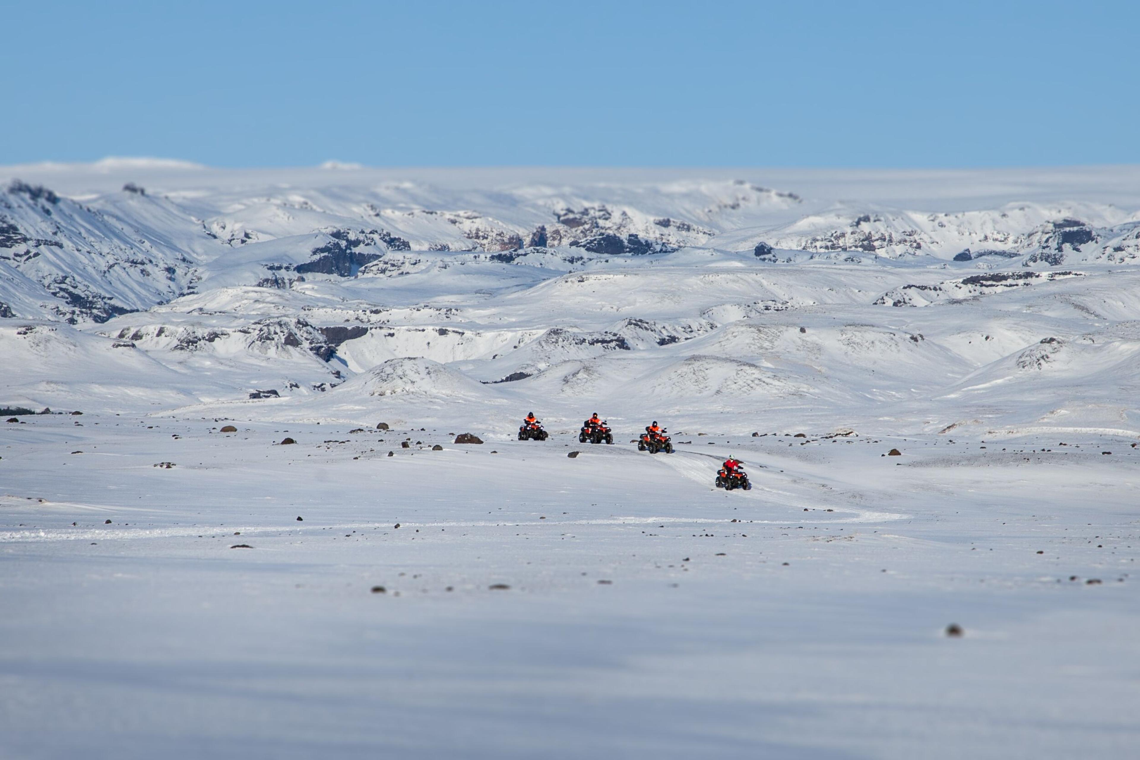 A group of ATV riders in bright orange gear traverse a vast, snow-covered landscape, with rugged, icy mountains stretching across the horizon under a clear blue sky.