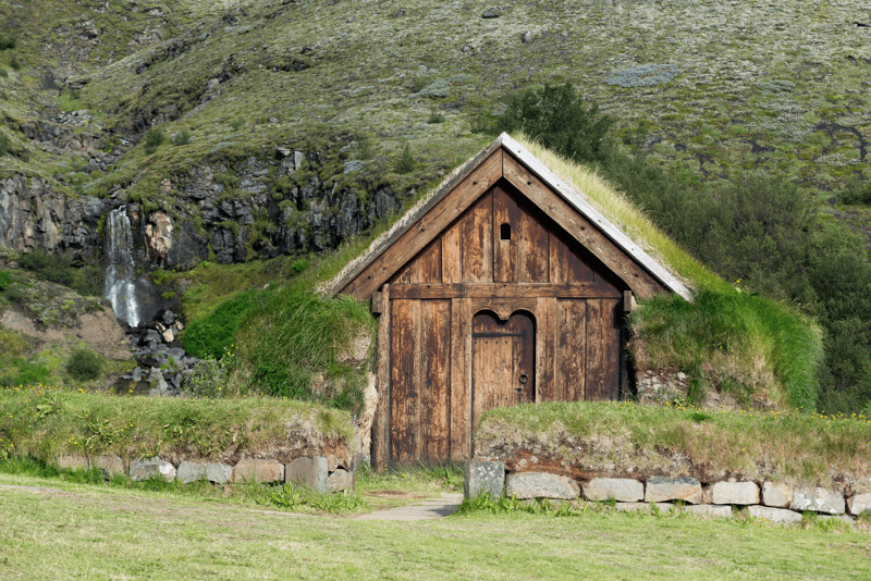 A traditional wooden turf house with a grass roof, a waterfall, and green hillside behind it.