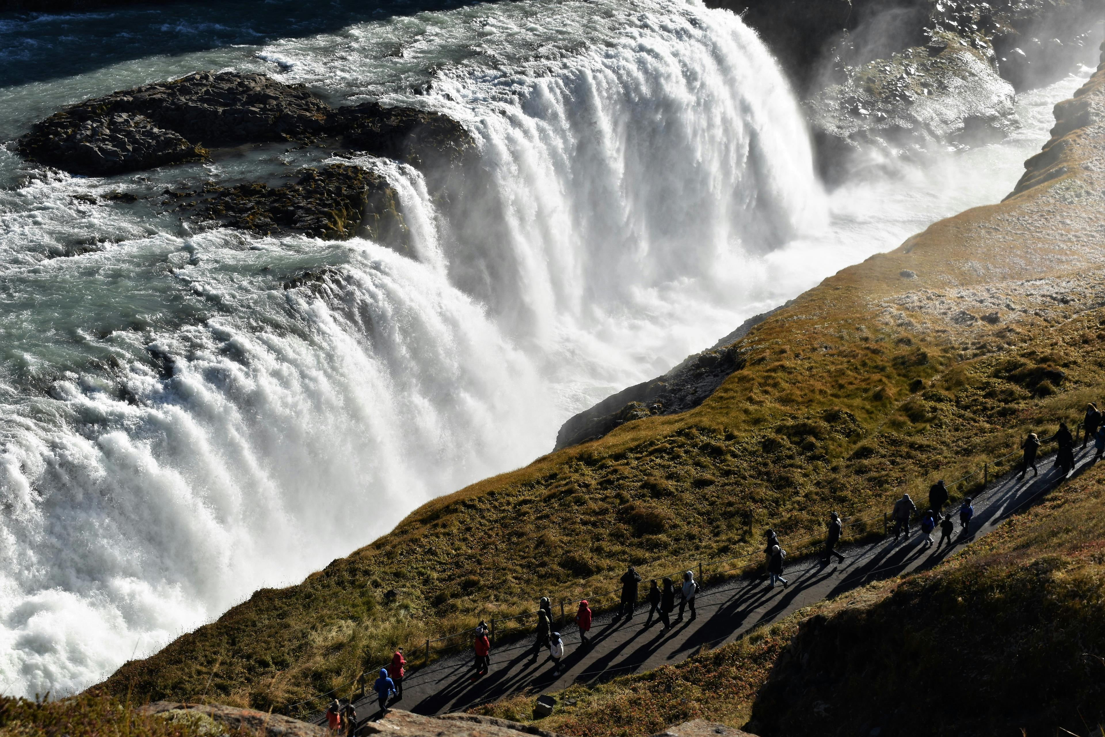 Visitors on a path observing a robust waterfall as it descends in a powerful rush, with the sunlight accentuating the spray and textures of the landscape, a scene likely from one of Iceland's popular natural attractions.