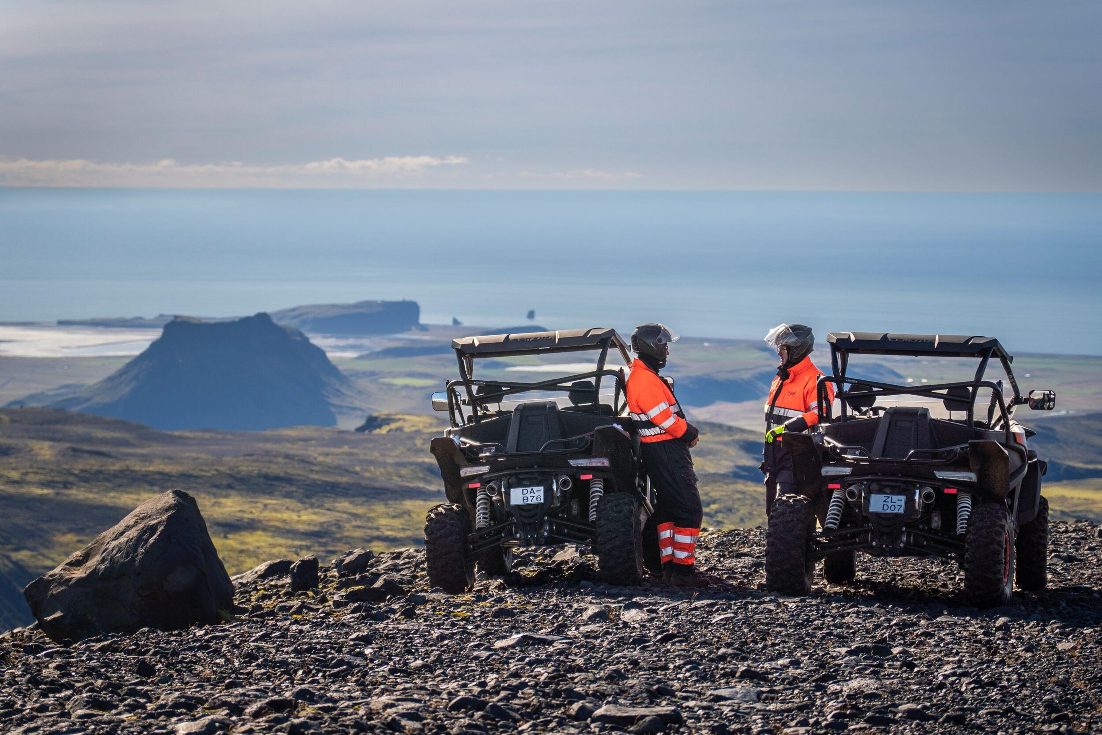 Two ATV riders in bright orange gear stand beside their vehicles on a rocky hilltop, gazing at the breathtaking coastal view of Iceland’s rugged landscape below.