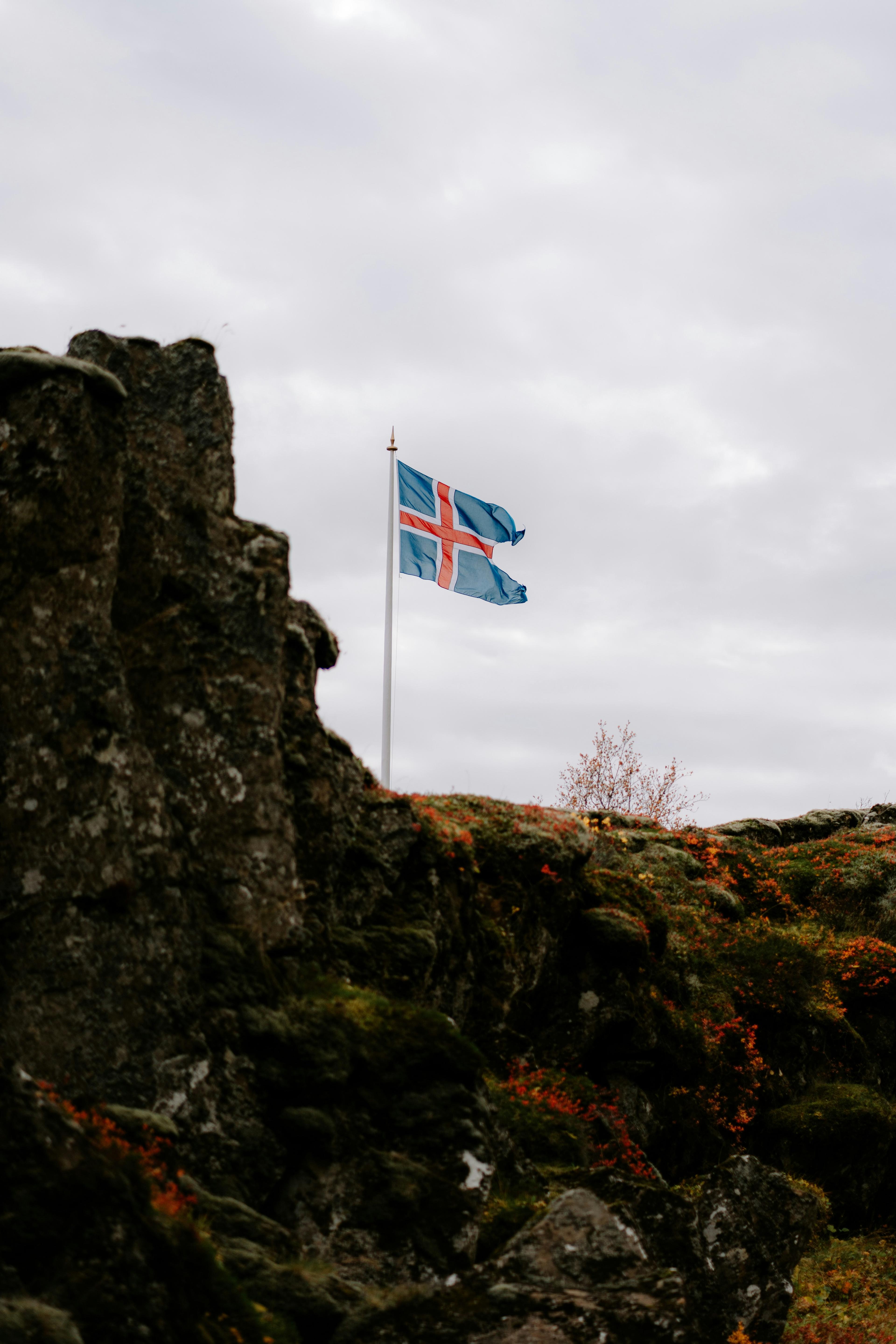 Icelandic flag waving atop "Lügverg", a moss-covered rocky hill under a cloudy sky, symbolizing national pride.